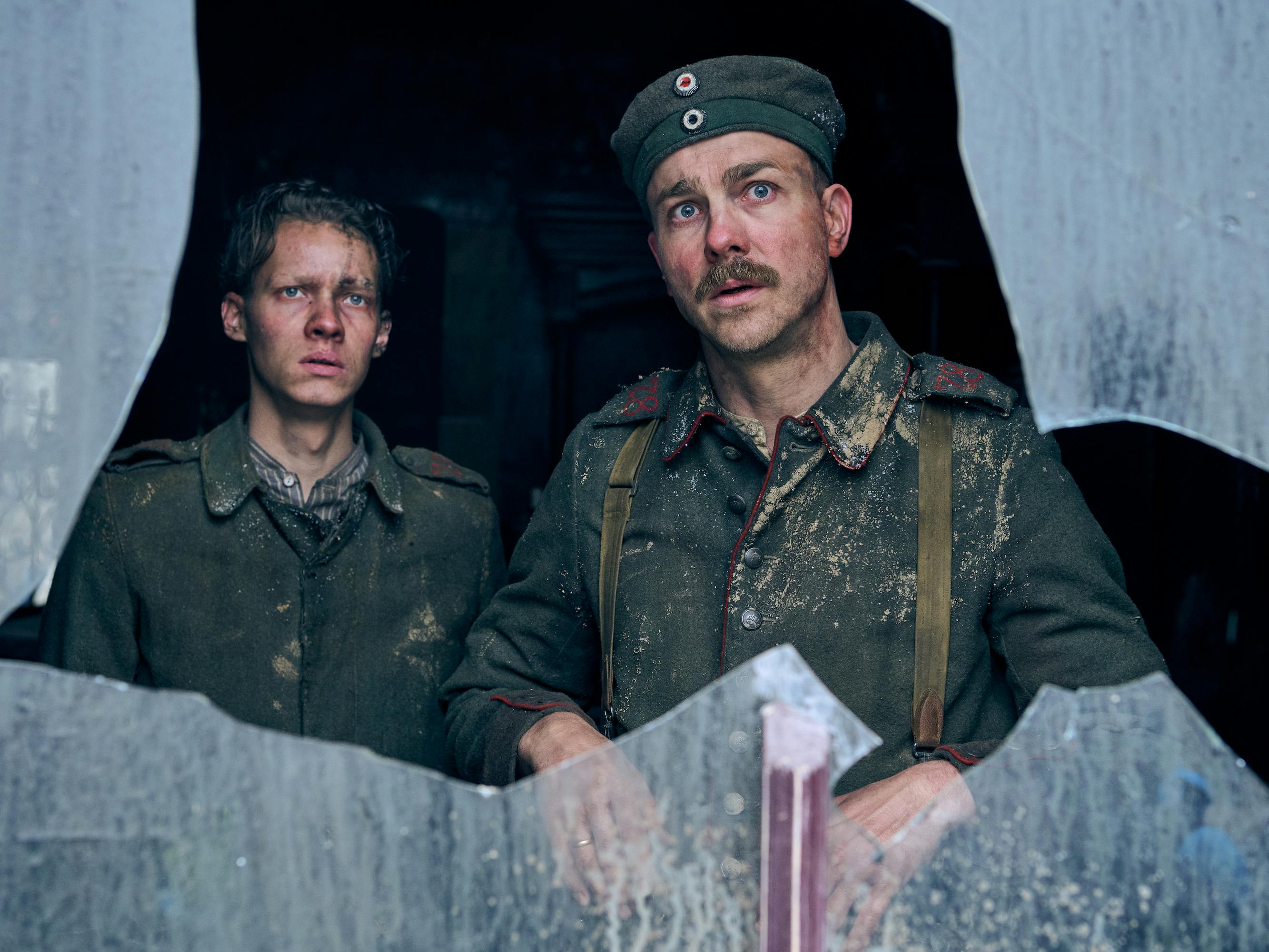 Paul Bäumer (Felix Kammerer) and Stanislaus Katczinsky (Albrecht Schuch) stand together behind a broken window in their green uniforms dusted with sawdust.