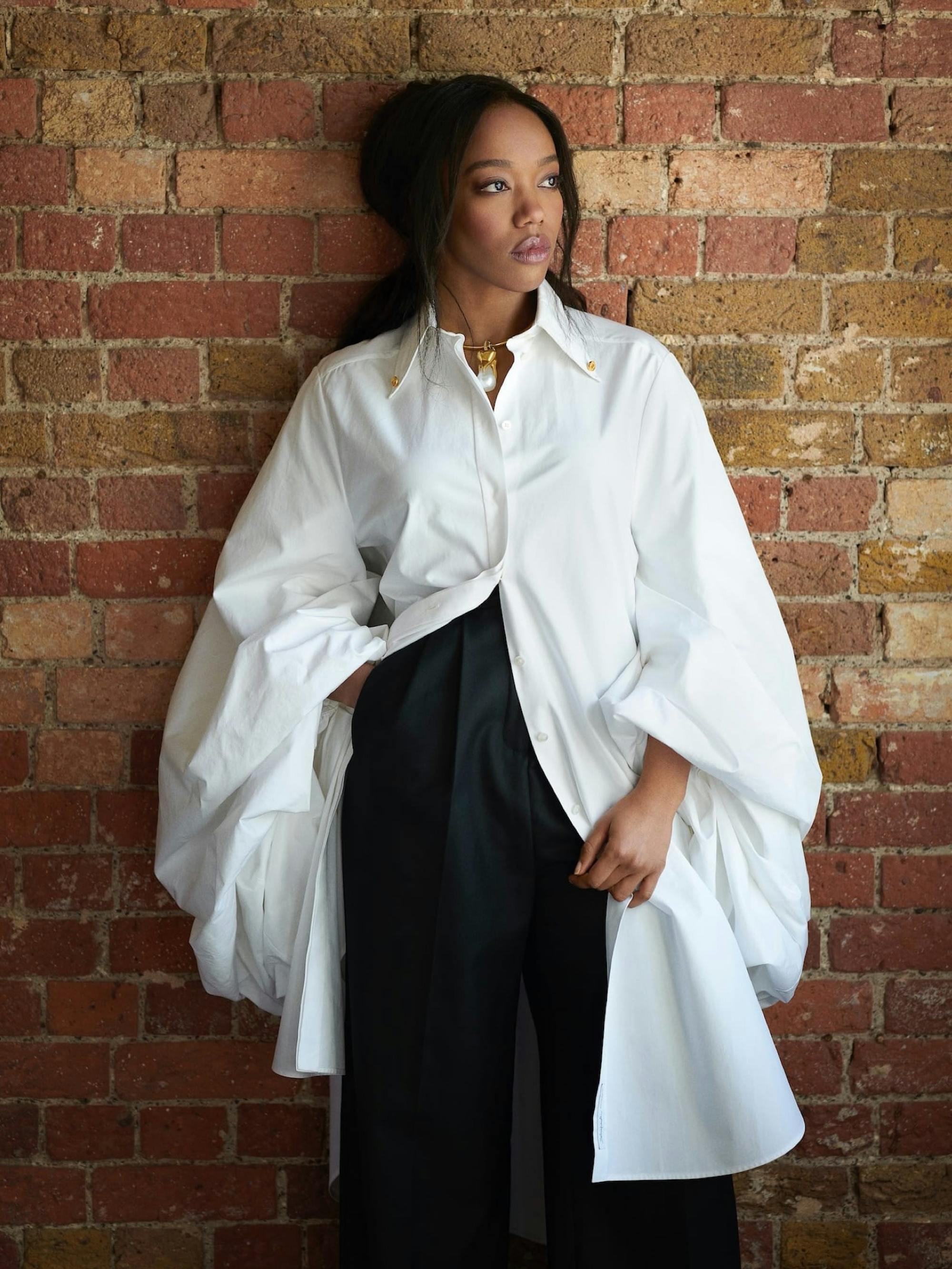 Naomi Ackie sports a long white buttoned-down shirt, with very wide sleeves, highwaisted black pants, and a fabulous gold and pearl necklace. She rests against a brick wall and looks into the distance.