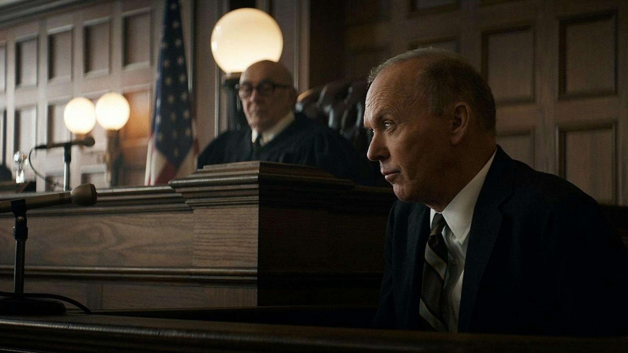 Judge Julius Hoffman (Frank Langella) and Ramsey Clark (Michael Keaton) in The Trial of the Chicago 7 sit behind the stand in a wood paneled courtroom wearing suits.