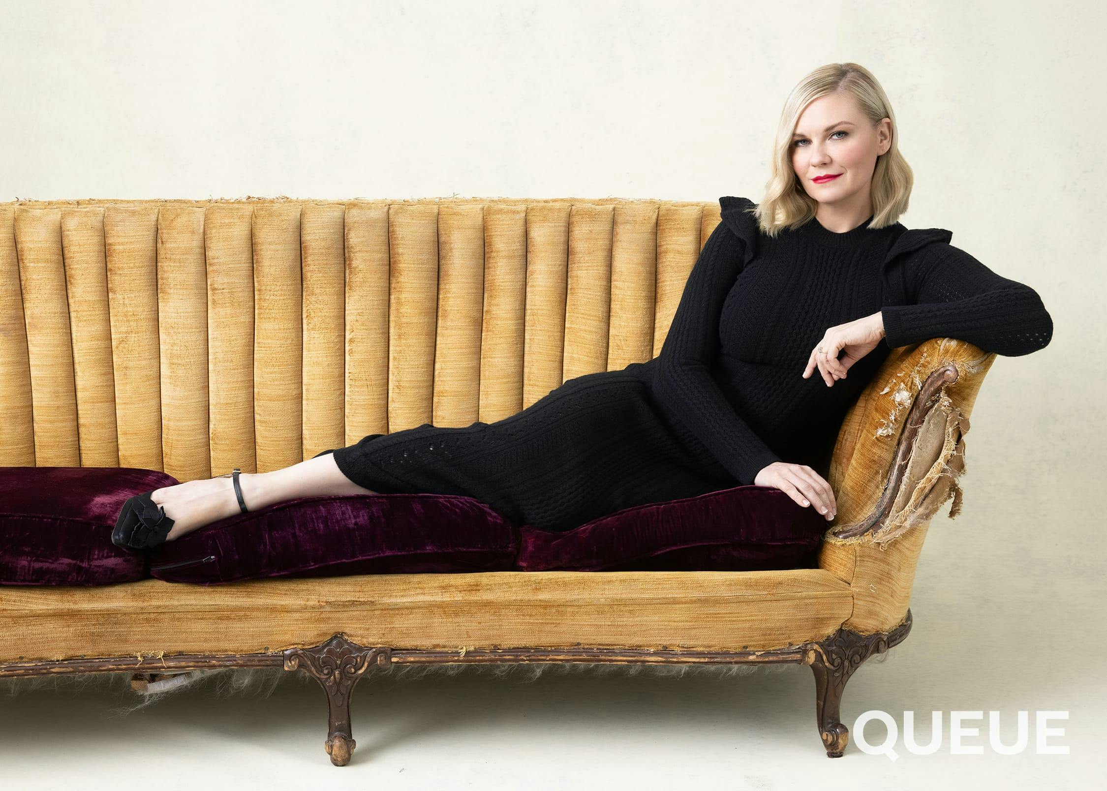 Kirsten Dunst wears a black dress and lounges on a beige and red couch.
