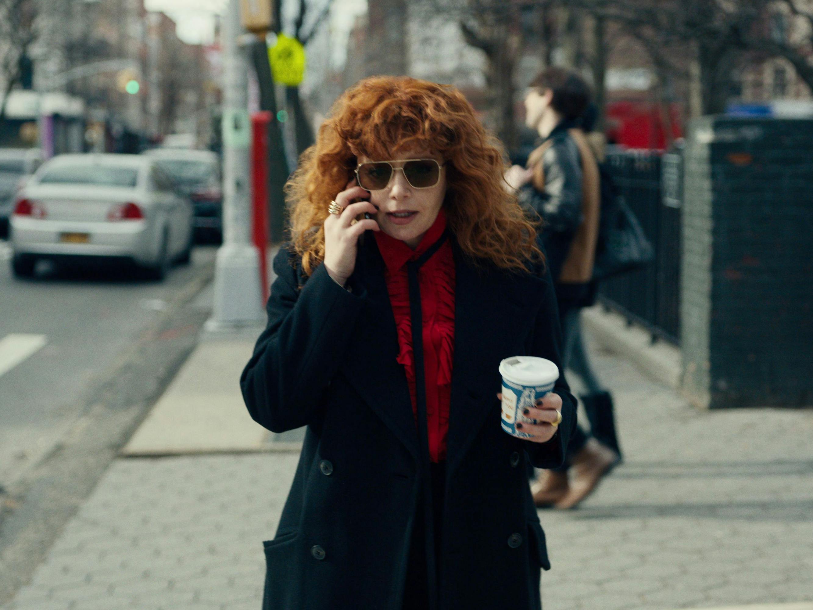 Natasha Lyonne wears a black coat, sunglasses, a red scarf and carries a coffee cup. She talks on the phone as she walks down the street.