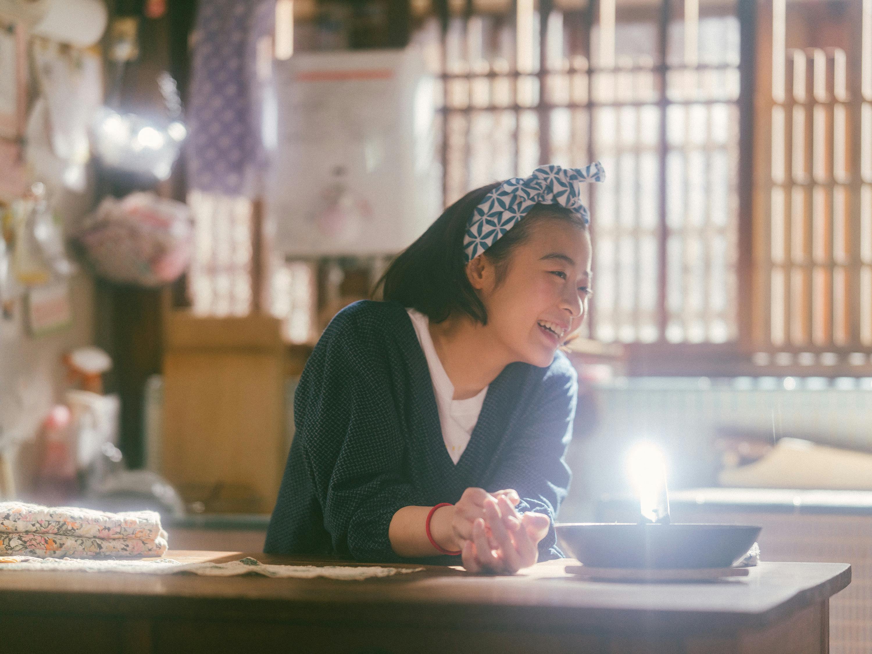 Kiyo (Nana Mori) smiles. She wears a patterned headband, white shirt, and a blue v-neck sweater.