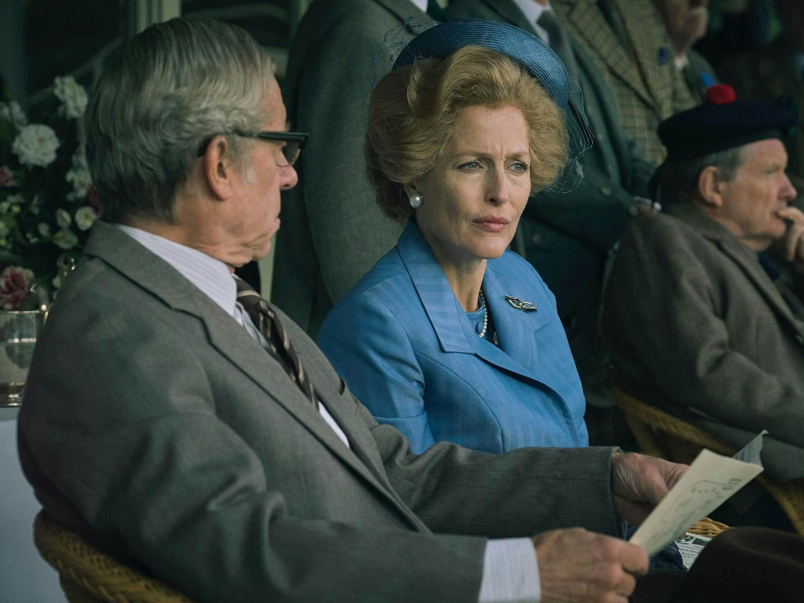 Denis Thatcher (Stephen Boxer) and Margaret Thatcher (Gillian Anderson) sit together. Denis wears a grey suit, and Margaret wears a blue suit and matching hat.