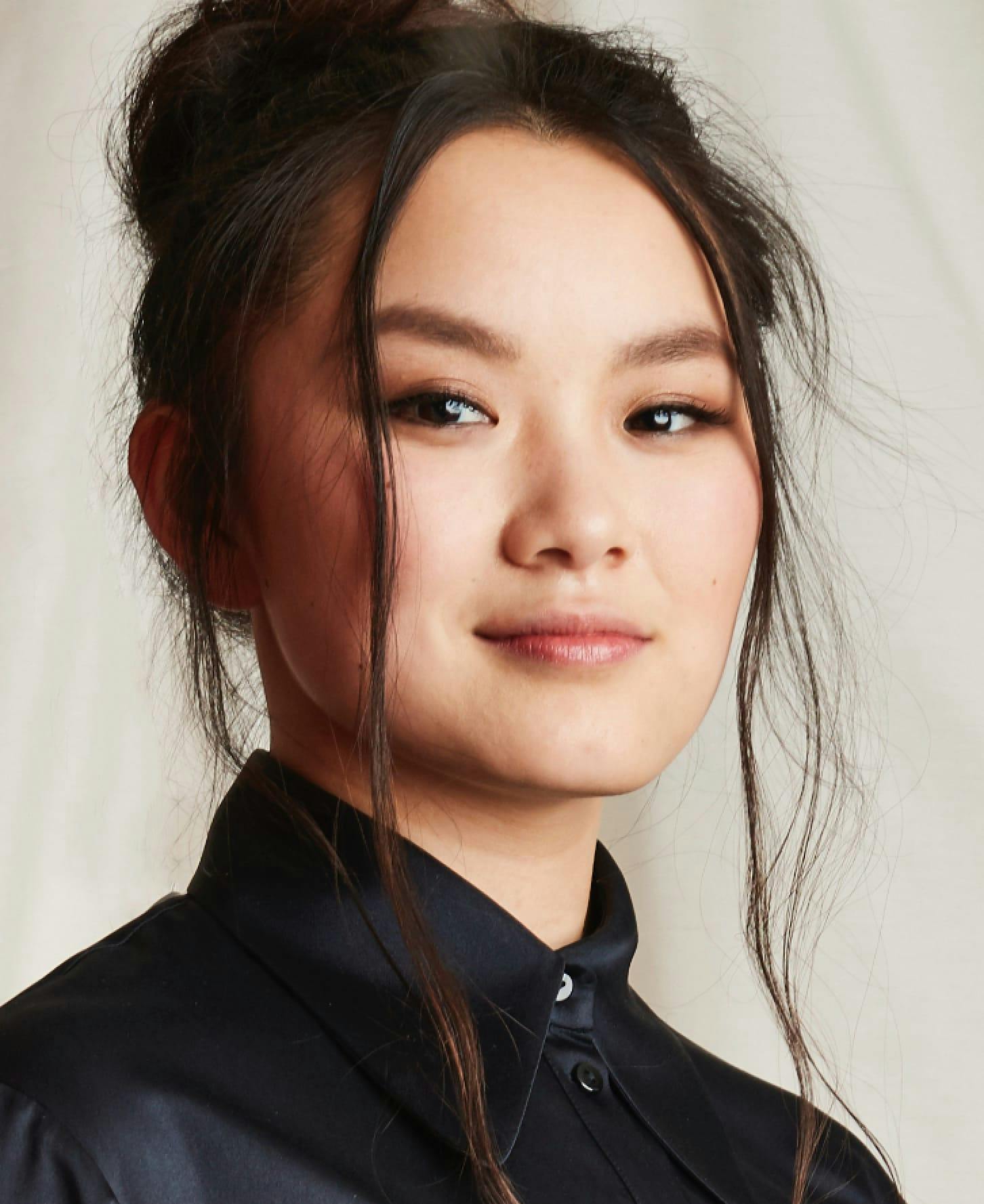 A portrait of Thaddea Graham. The actor is pictured from the shoulders up, wearing a black silk shirt buttoned to the neck. Her hair is up in a bun with two long strands framing her face. She turns her head slightly to look at the camera with a confident gaze. 