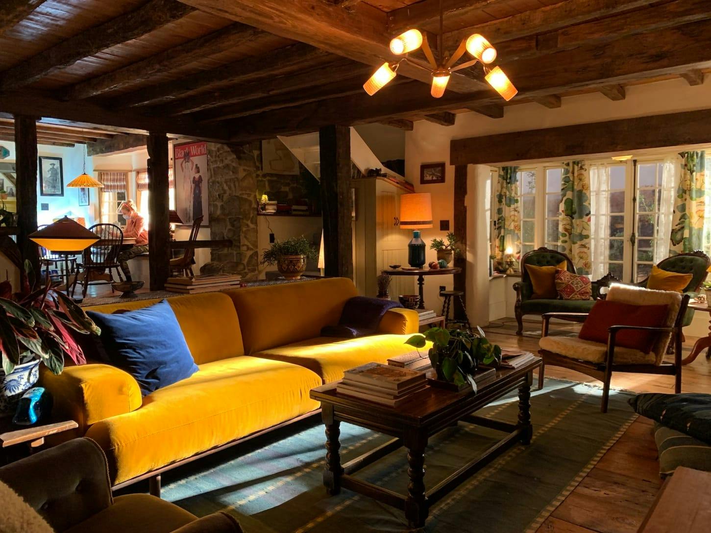 The living room. There is a yellow velvet couch, coffee table with books and a plant, overhead light fixture, chairs, more plants, pillows, and french doors in the background.