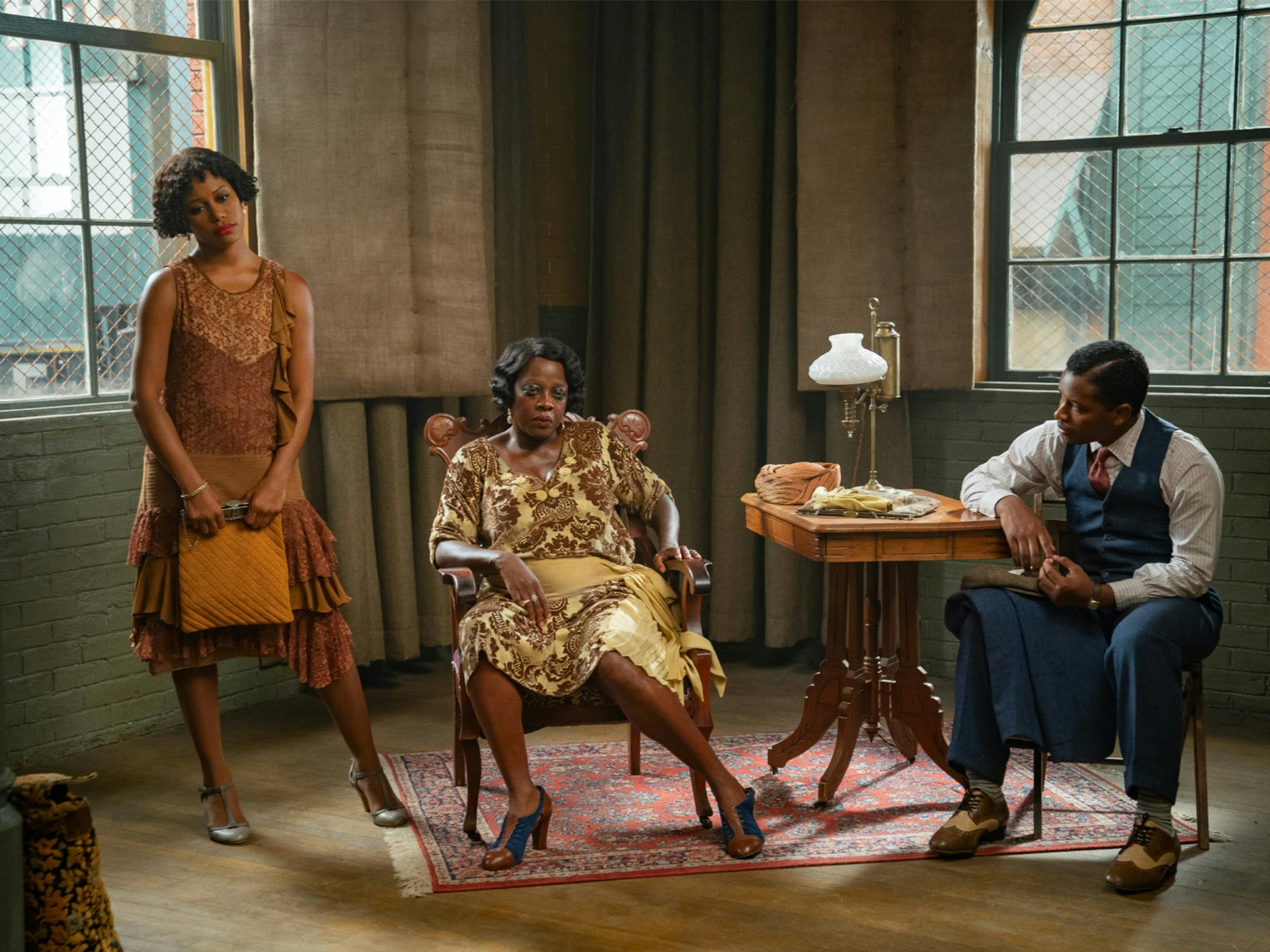Ma Rainey slumps casually in an armchair at the recording studio. Her lover Dussie Mae stands to her right, leaning her weight on one hip and looking bored. Her nephew Sylvester is in a chair to her left, looking over at her. Greens, browns, and blues pervade the scene.