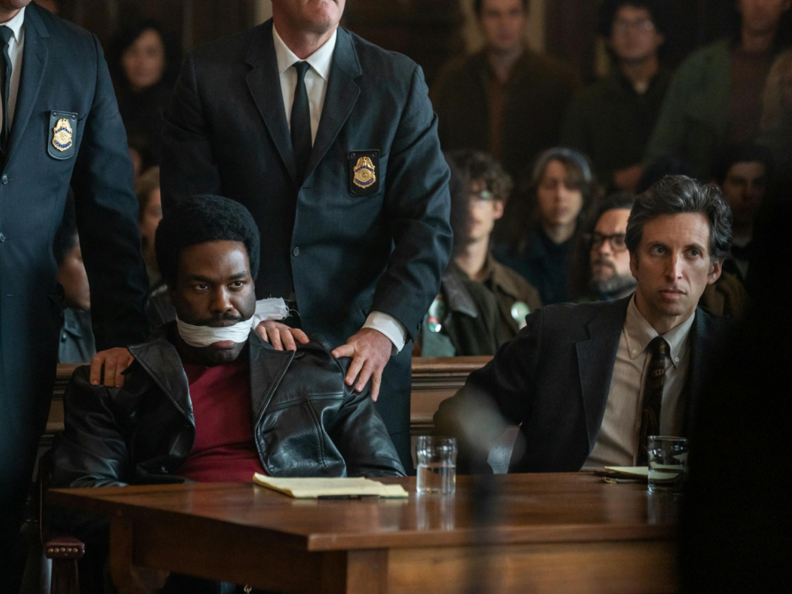 Abdul-Mateen in a scene from The Trial of the Chicago 7. He portrays Bobby Seale bound and gagged. Two bailiffs placing hands on his shoulders. He looks willfully toward the bench.