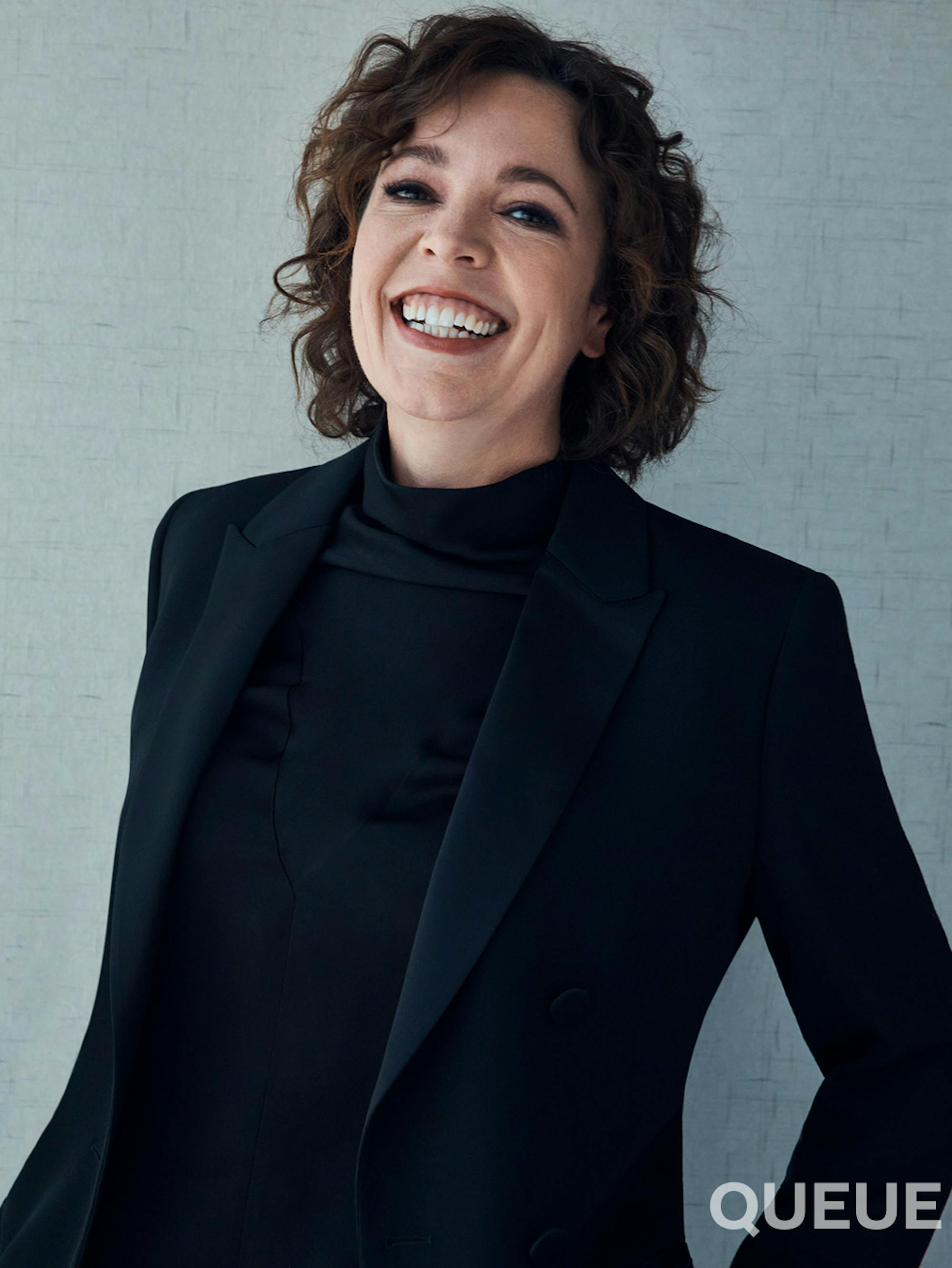  Olivia Colman wears a dark blazer and turtleneck as she smiles wide.
