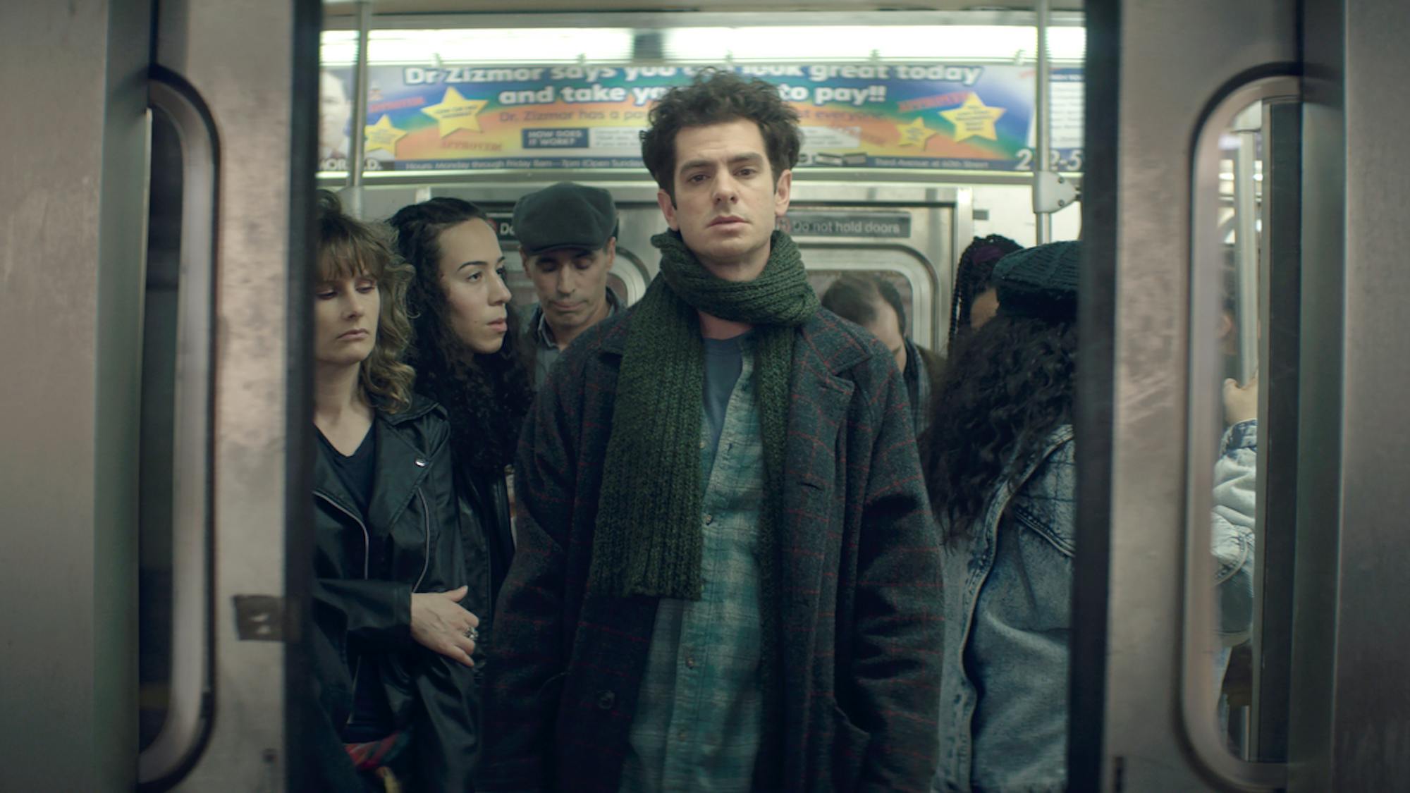 Andrew Garfield as Jonathan Larson wears a dark jacket, checkered shirt, and green scarf as he stands between closing subway doors.