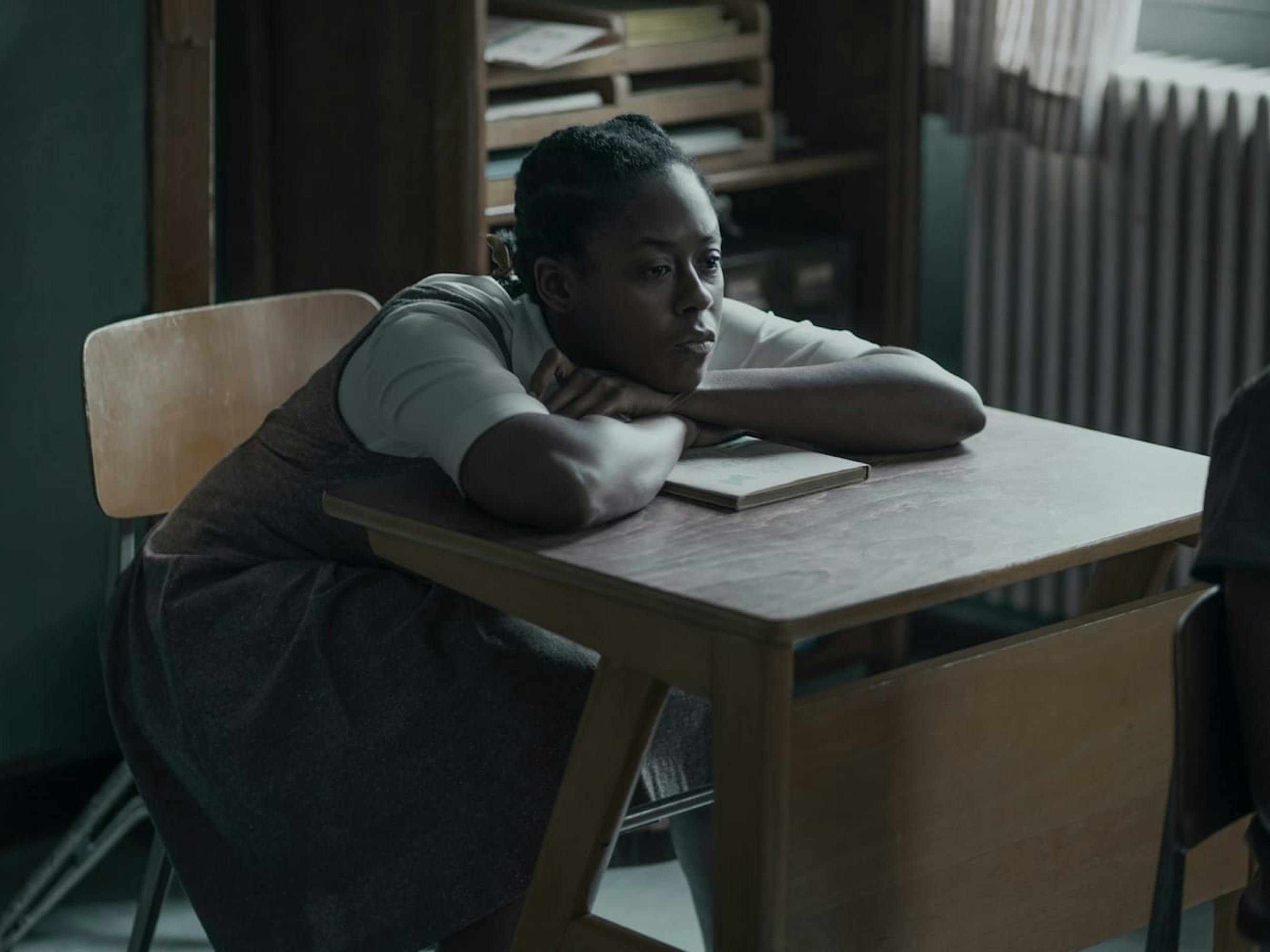 Jolene (Moses Ingram) rests against a desk in a classroom. The desk and chair are wooden, and there are books in a shelf behind her. She wears a long dark dress with a white shirt beneath, and looks bored. 