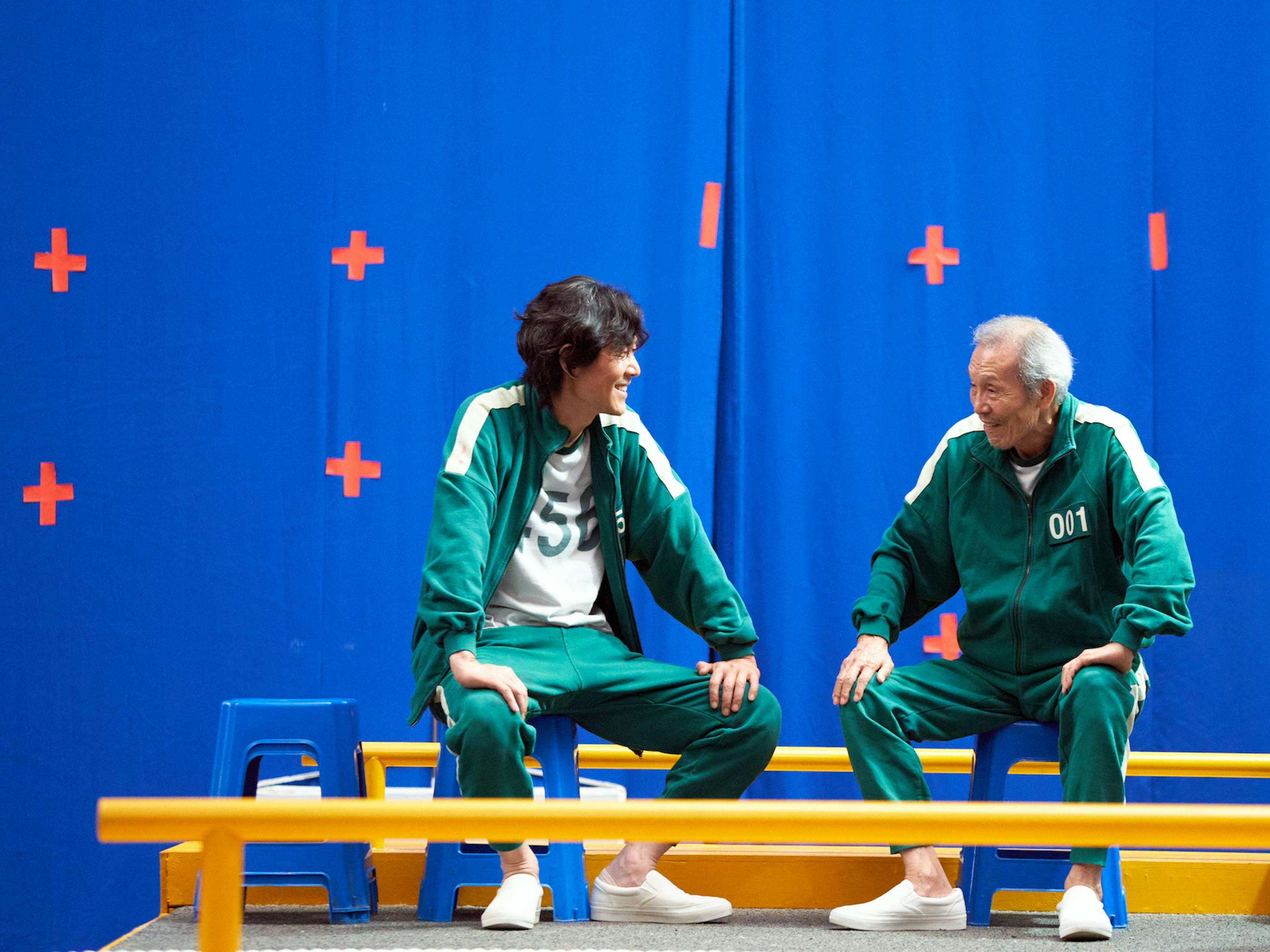 Seong Gi-hun (Lee Jung-jae) and Oh Il-nam (Oh Young-soo) sit on a yellow bench in their green tracksuits.