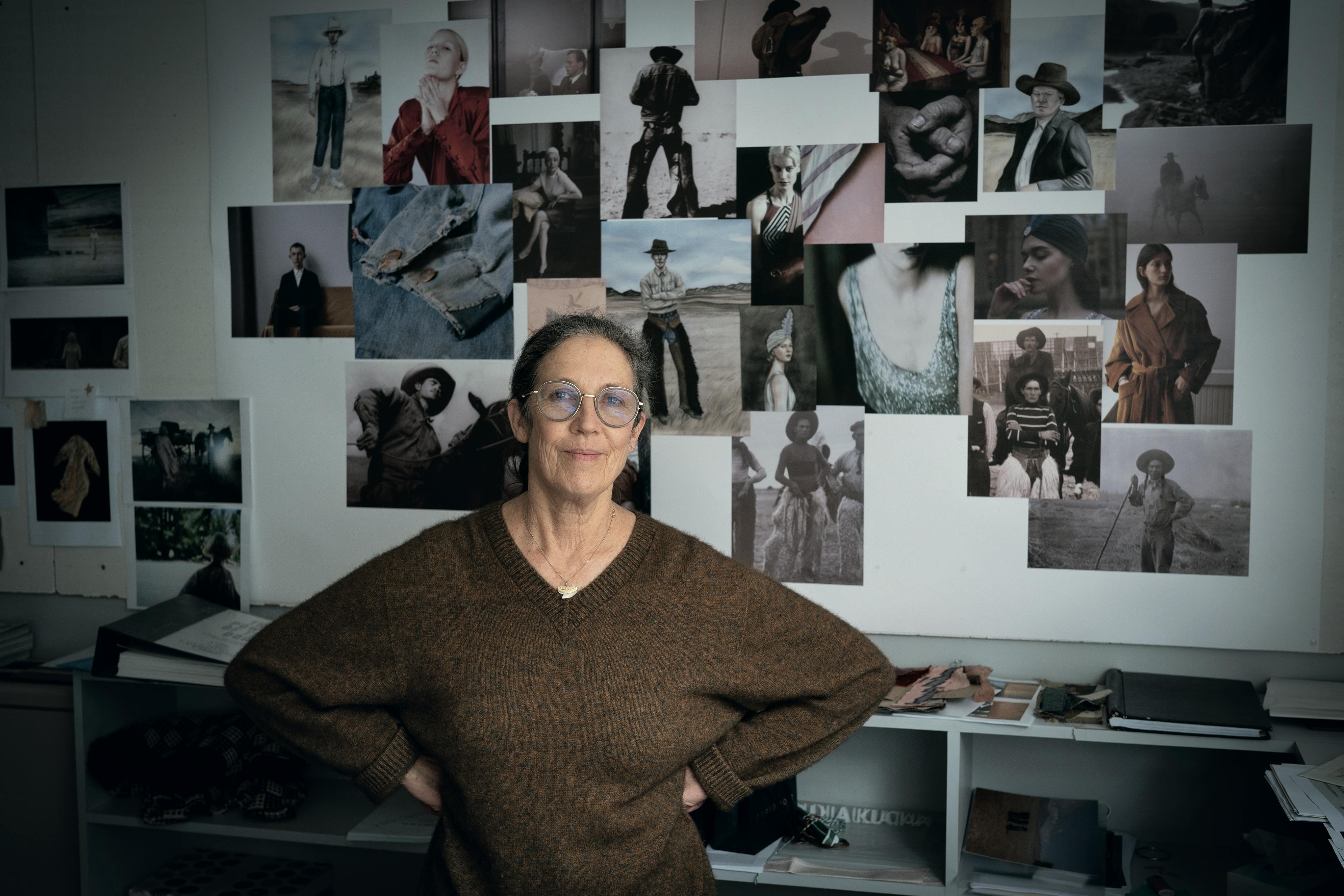 Kirsty Cameron wears a brown sweater and stands in front of a wall plastered with photos.