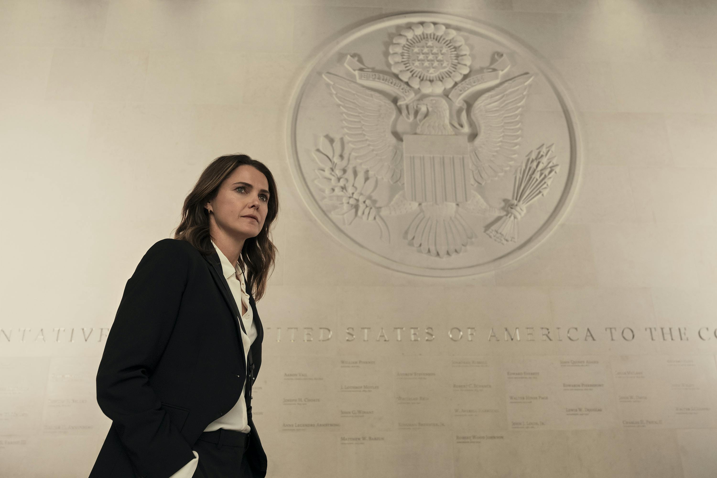 Kate Wyler (Keri Russell) walks past a U.S. seal. She wears a black suit and a white shirt and looks stressed. 