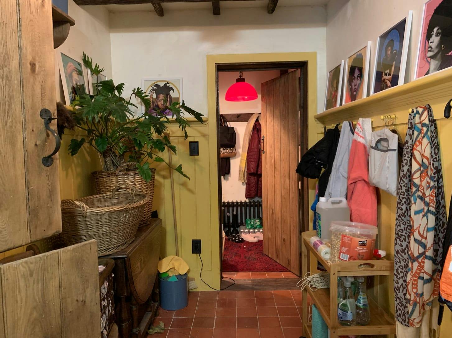 The mudroom, finished with bright yellow. There are portraits against the wall, hanging clothes, a basket, and a huge plant. There is also a closet that is lit by a red overhead light.