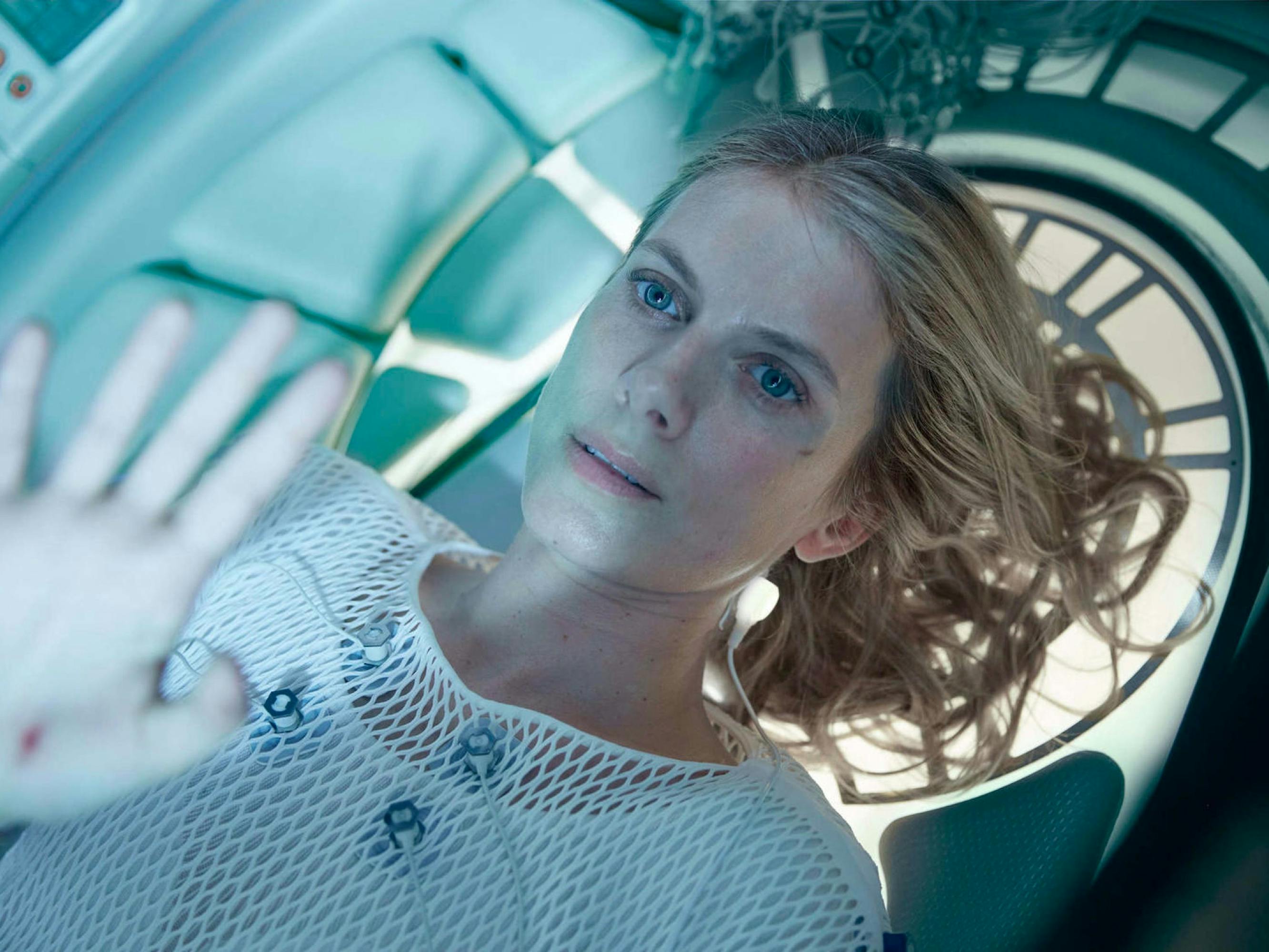 Laurent lies in her cryogenic chamber, hand pressed against the top giving a sense of how small the space is. She wears a white mesh top with some medical cords attached to her front. The chamber resembles a spaceship.