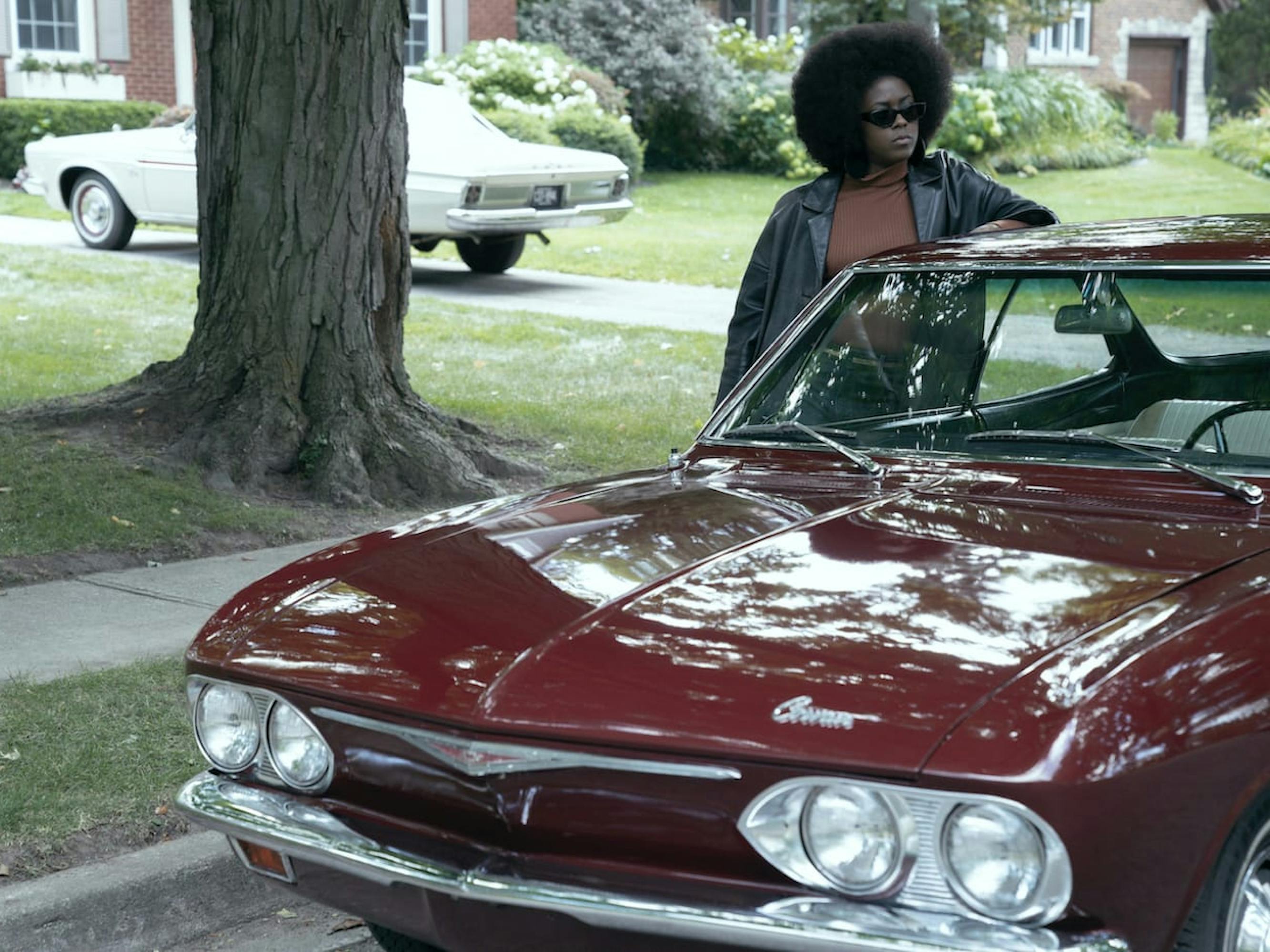 Jolene (Moses Ingram) rests against a red convertible, in a suburban neighborhood. She wears an orange shirt, black leather jacket, and black shades, looking overall very badass.