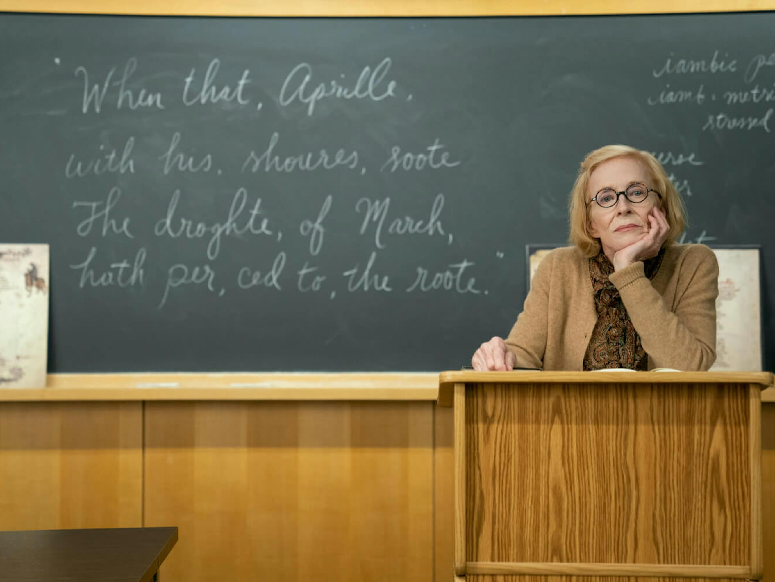 Professor Joan Hambling rests her chin on her hand looking bored. She stands behind a wood podium. In the background is a chalkboard with quotes written in white chalk.