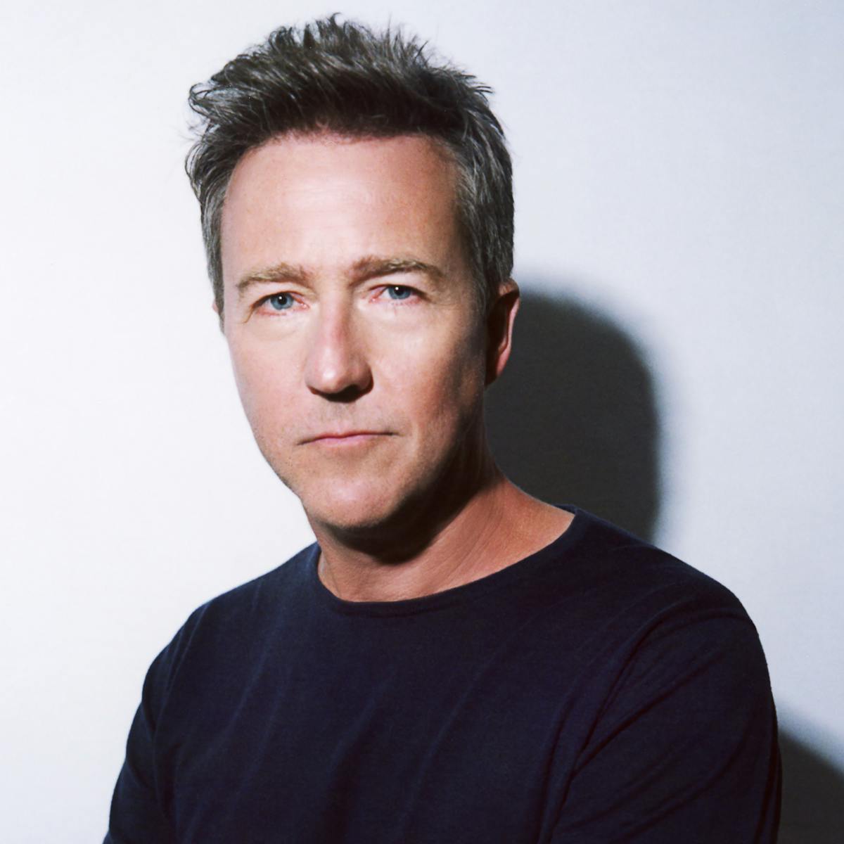 Edward Norton wears a dark shirt against a white background and smiles slightly.