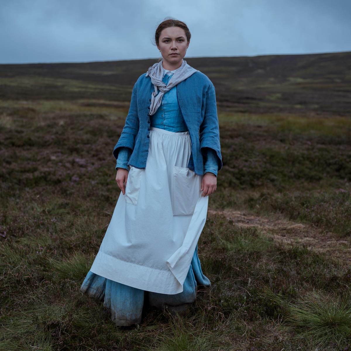 Nurse Lib (Florence Pugh) wears a blue dress and white apron and stands in a grassy field.