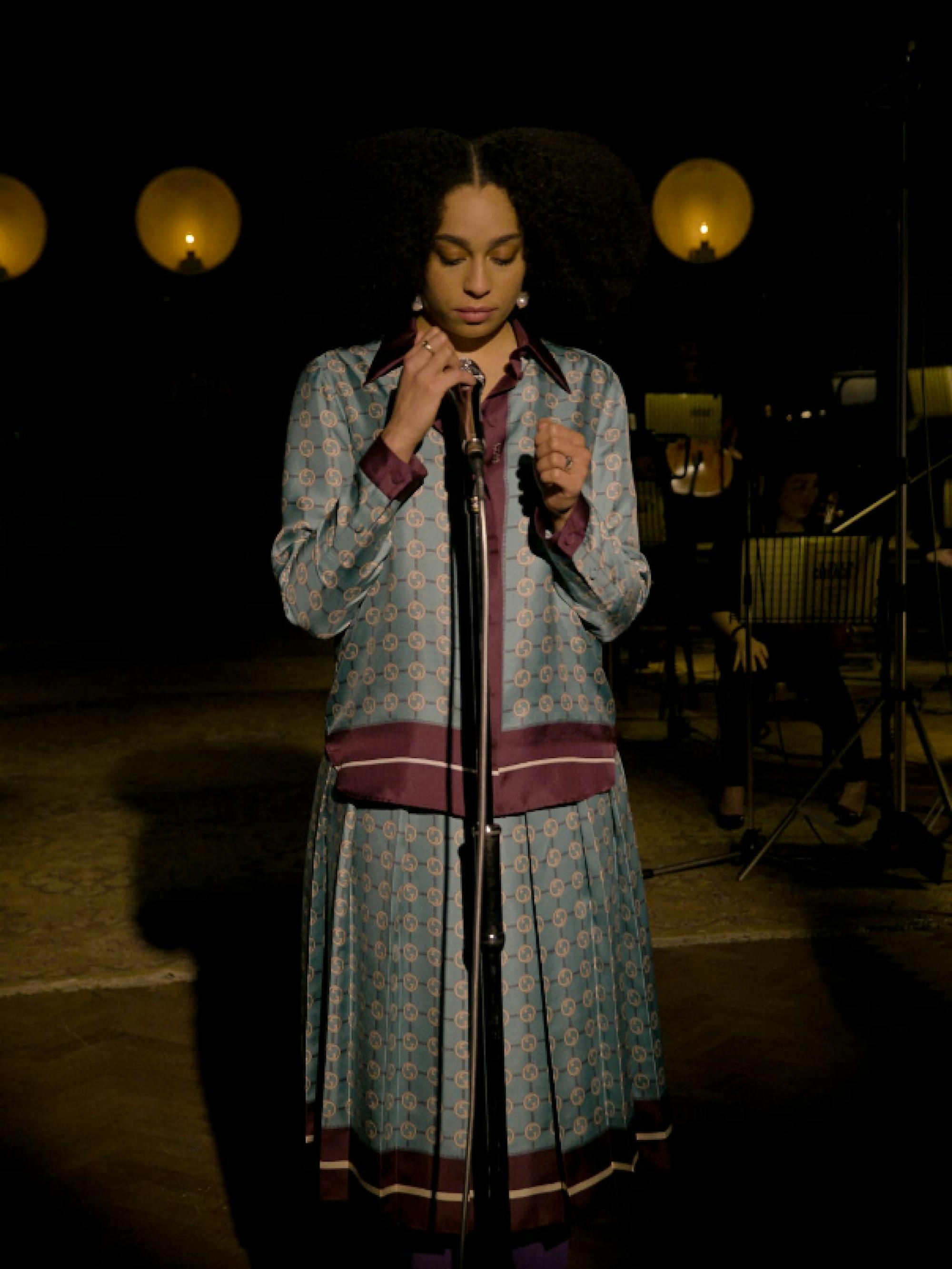 Singer Celeste performs her song at a microphone stand. Dim lights are lit behind her. She’s wearing a gorgeous blue and purple dress that feels like it could have fit in during the era of the film for which she wrote her song, “Hear My Voice.”