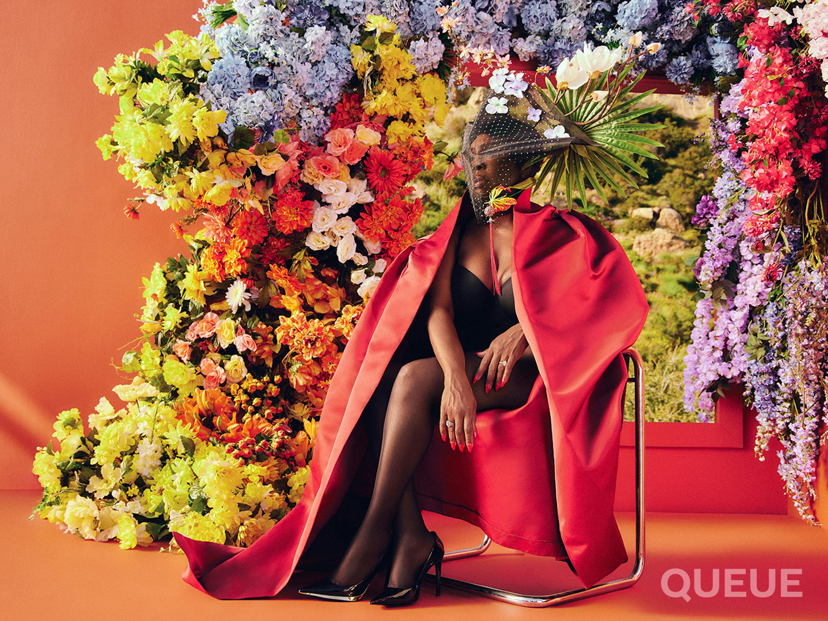 Niecy Nash-Betts sits on a chair wearing a black bodysuit and a red cape. She also wears a veil that has a few white flowers on it and a fanned-out palm frond. Behind her is a doorway covered in flowers.