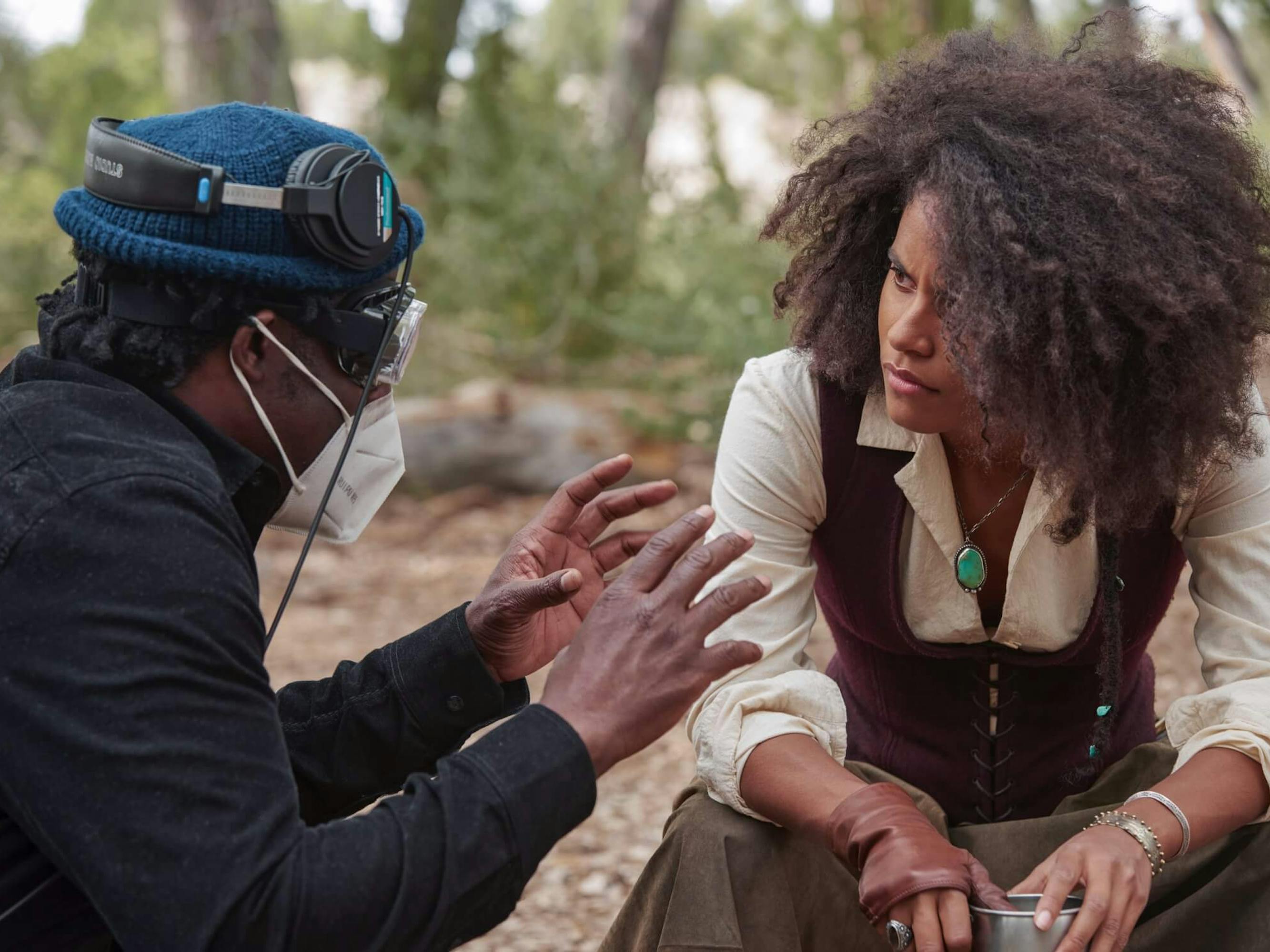 Jeymes Samuel and Zazie Beetz sit together. Samuel, in all black and a teal beanie, explains something to Beetz, who wears khaki pants, a white blouse, turquoise necklace, and brown vest.