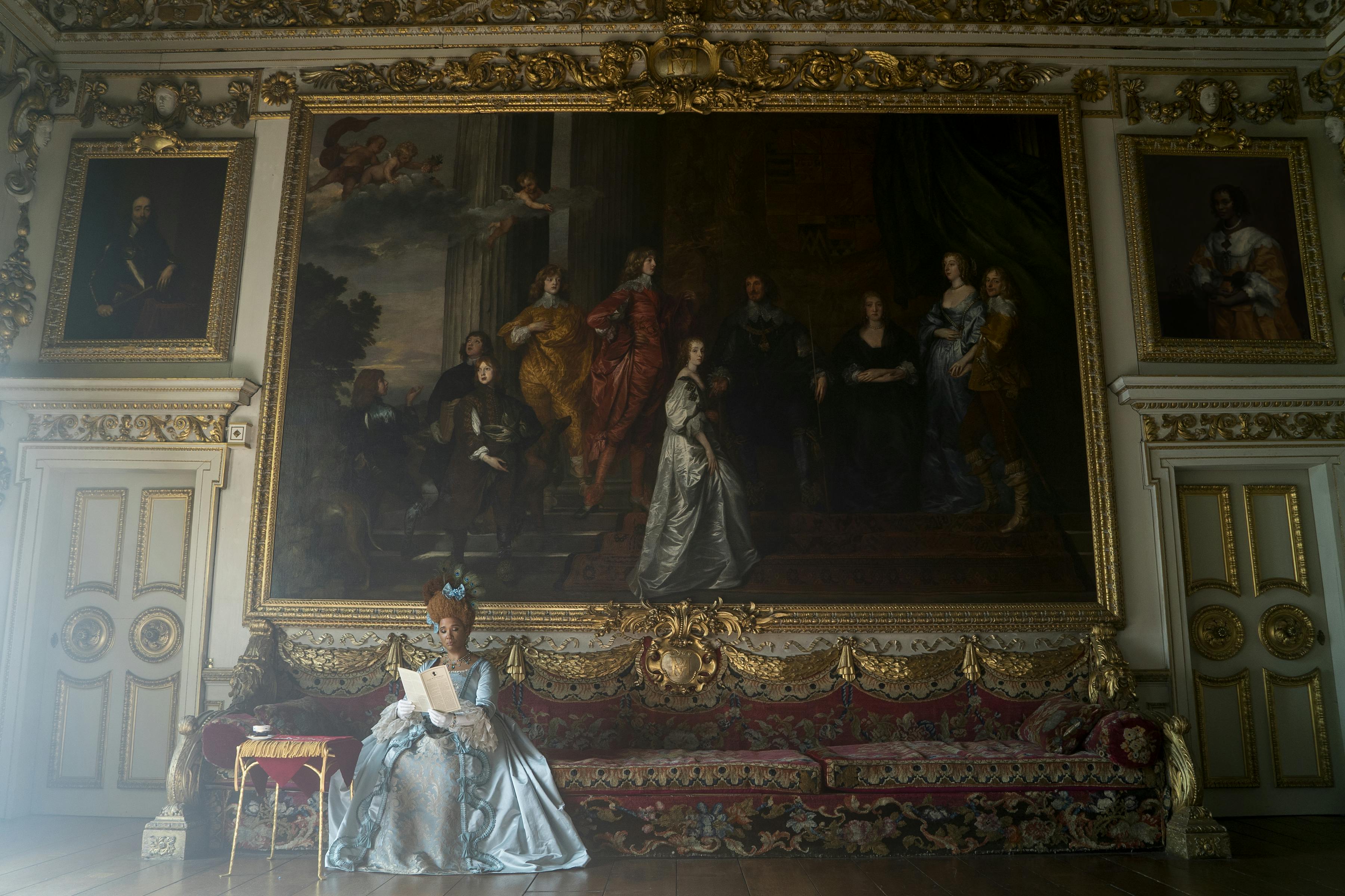 The Queen rests on an ornate couch patterned with gold trimming and red patterns. She wears a blue dress and blue headpiece. Behind her is a massive painting. 