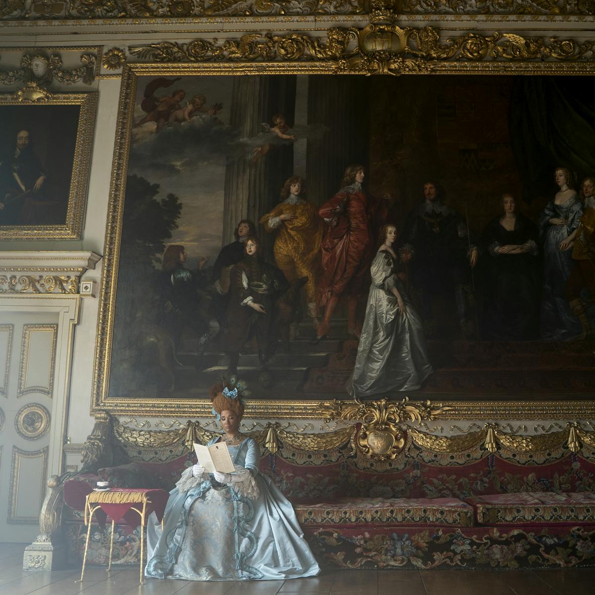 The Queen rests on an ornate couch patterned with gold trimming and red patterns. She wears a blue dress and blue headpiece. Behind her is a massive painting. 
