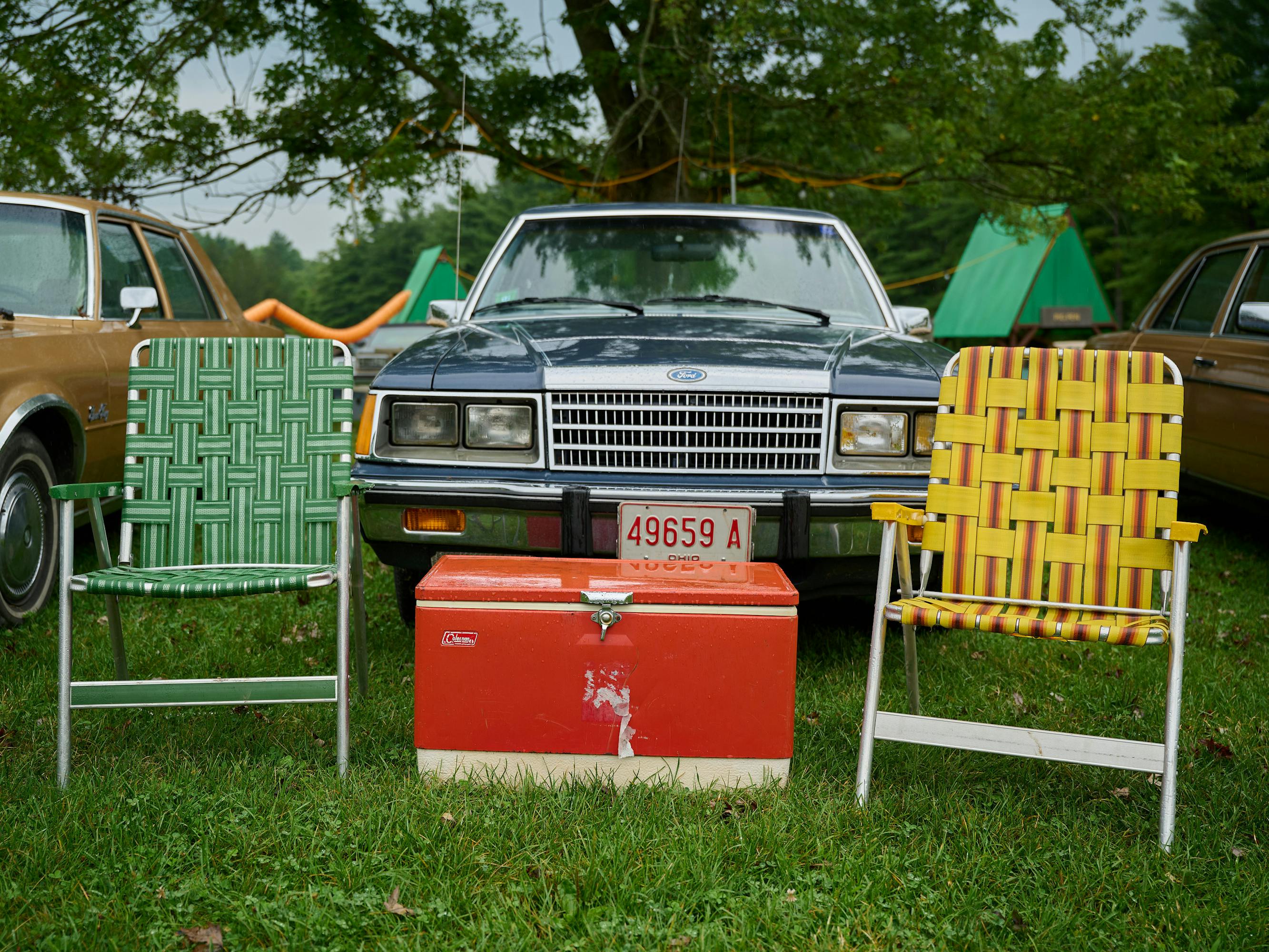 A cooler sits between a yellow and a green lawn chair on a grassy parking lot. A station wagon is behind them.