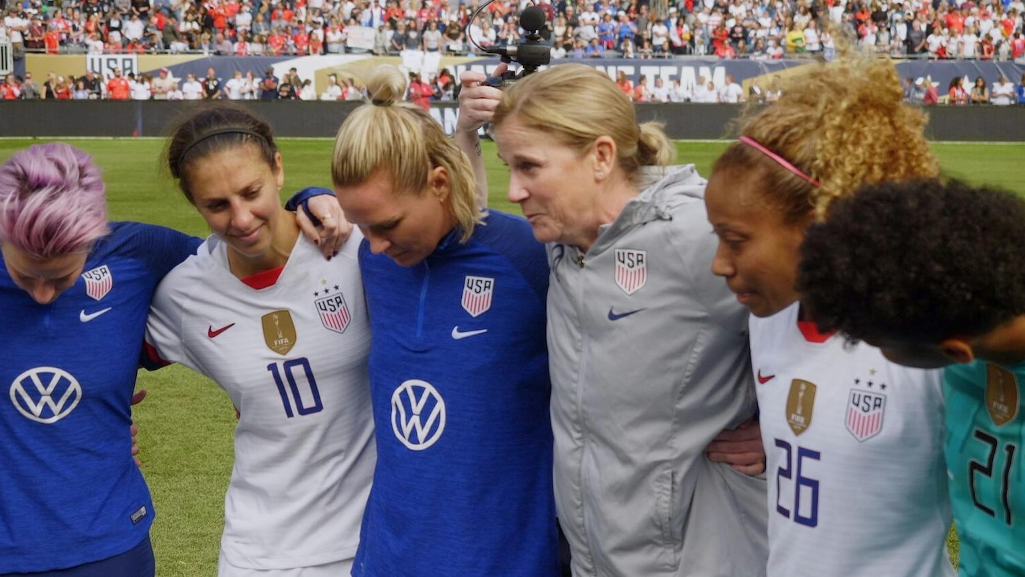 Megan Rapinoe, Carli Lloyd, Emily Sonnett, Jill Ellis, Casey Krueger, and Lynn Williams stand in a huddle on the soccer pitch. In the background are fans, and to their right is a camera. They wear blue and white jerseys except for the coach who wears a grey hoodie.