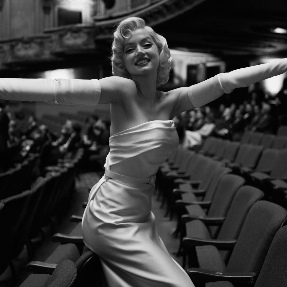 Marilyn monroe (ana de armas) wears a long white dress and stands in a empty theater with her arms outstretched.