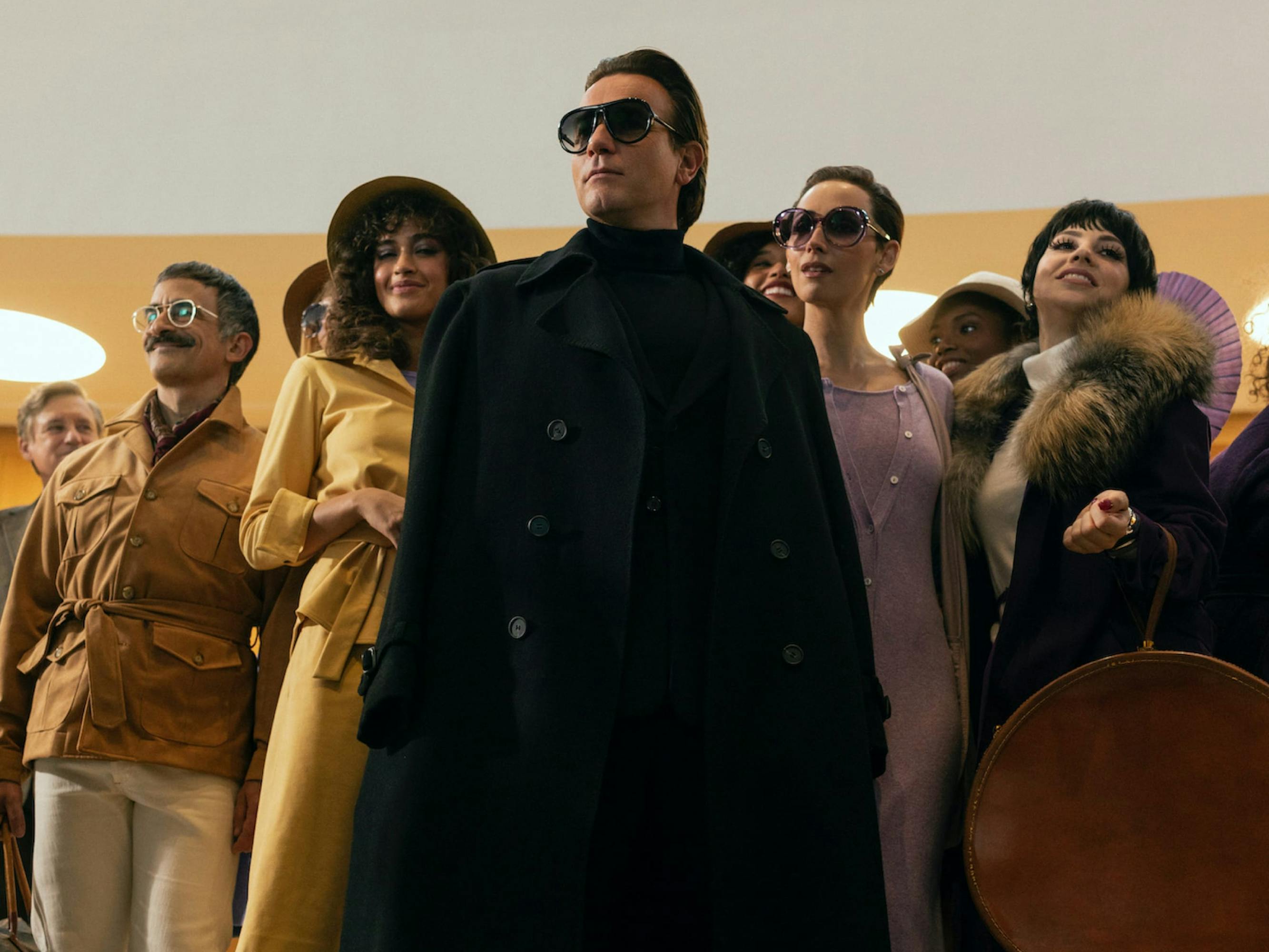 Halston (Ewan McGregor) wears a black double breasted coat, black turtleneck, black pants, and black glasses. Behind him stand an assortment of people wearing tans, purples, and blacks. Liza Minnelli (Krysta Rodriguez stands to his left, wearing a fabulous fur lined coat and carrying a brown circular bag. All of the people in this shot look off into the distance as if about to embark on an adventure.