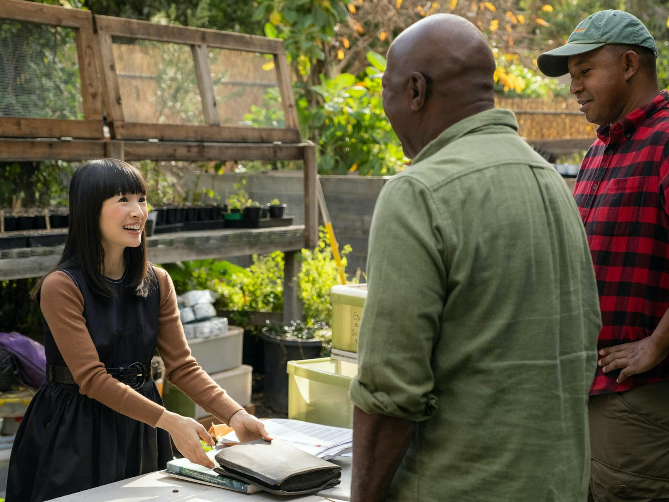Marie Kondo wears a belted dress and long sleeve shirt and stands opposite clients Logan and Jim, both wearing button-downs, outdoors at their plant nursery.