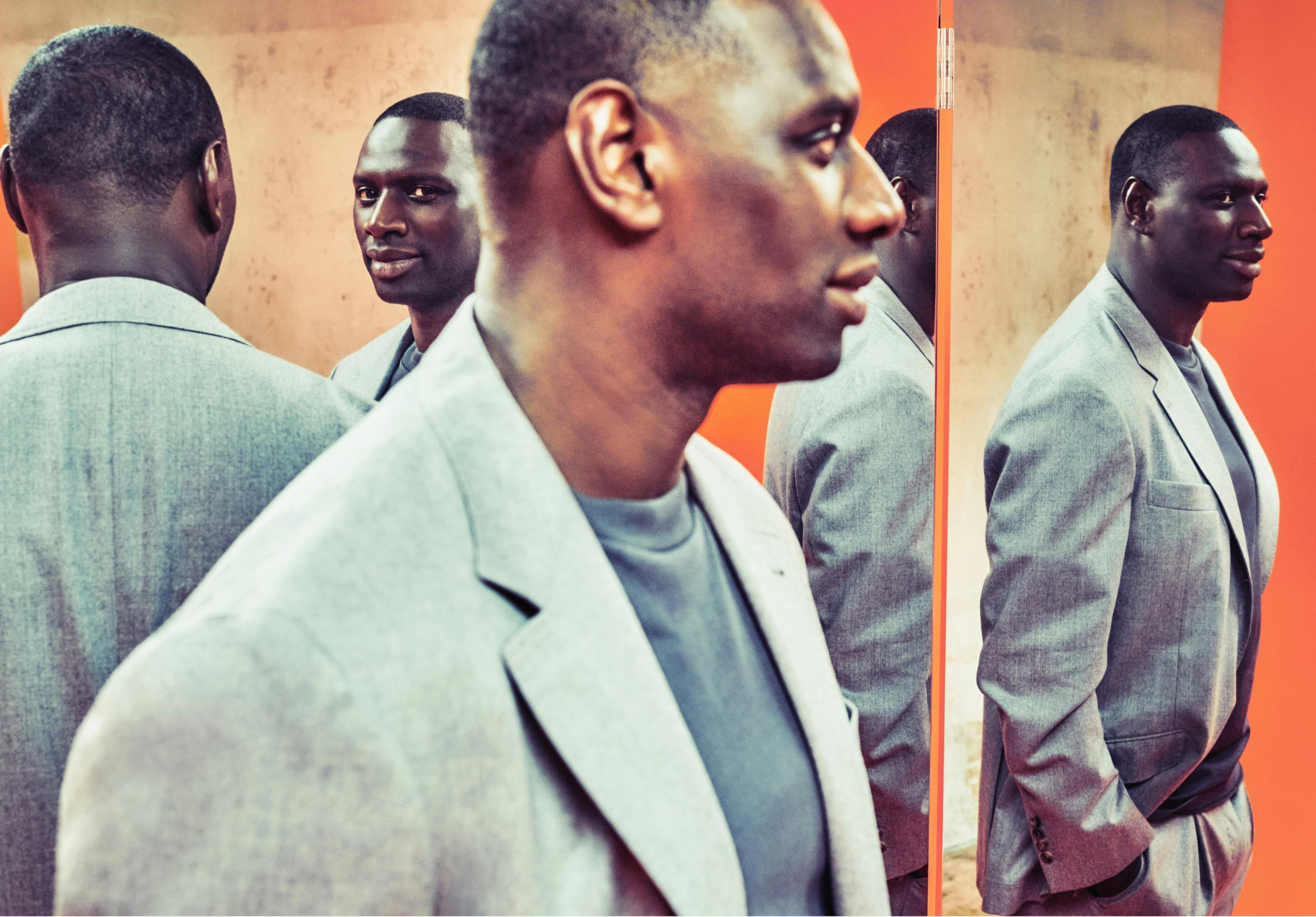 Reflections of Omar Sy populate this brightly colored image. The actor is pictured in a light-colored blazer, which pops against a strong orange background. 