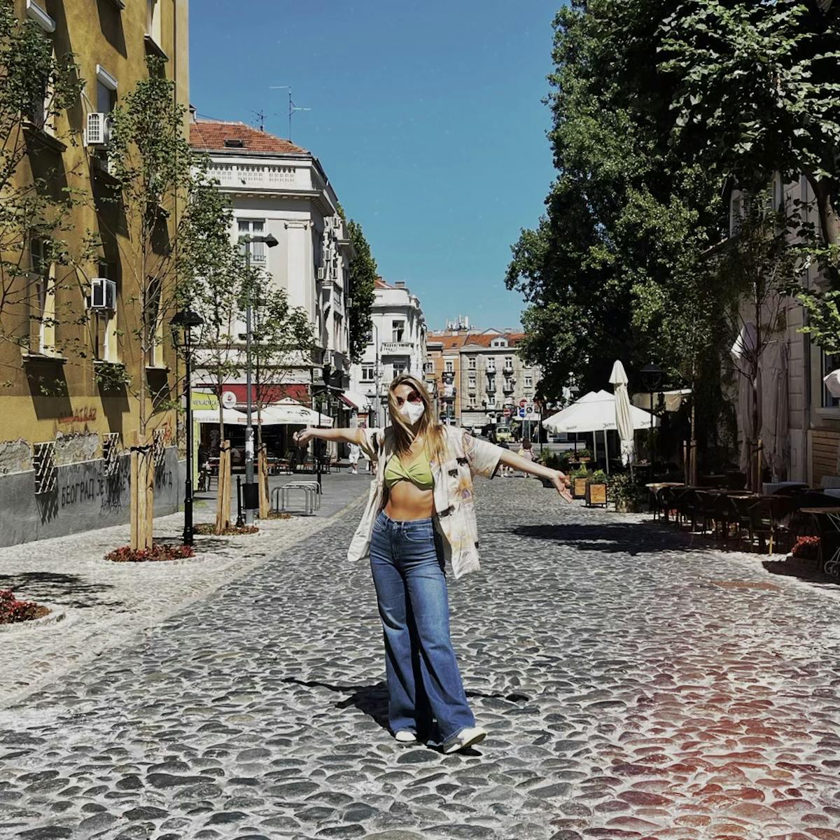 Madelyn Cline wears baggy blue jeans and a green crop top with a mask and sunglasses. She stands in a cobble street on a sunny day.