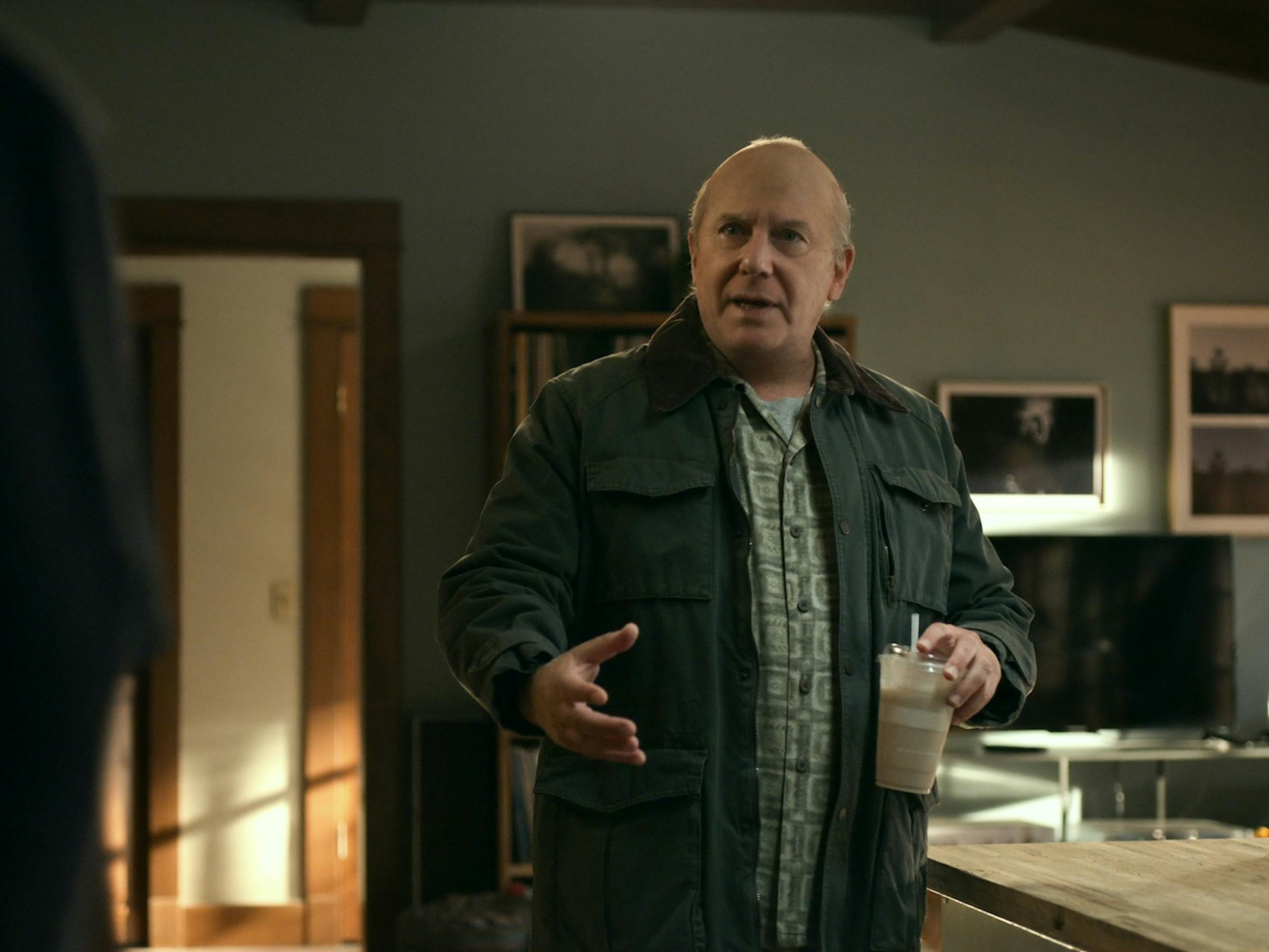 Martin (Paul Reiser) wears a green jacket, holds an iced coffee, and motions to someone out of view.