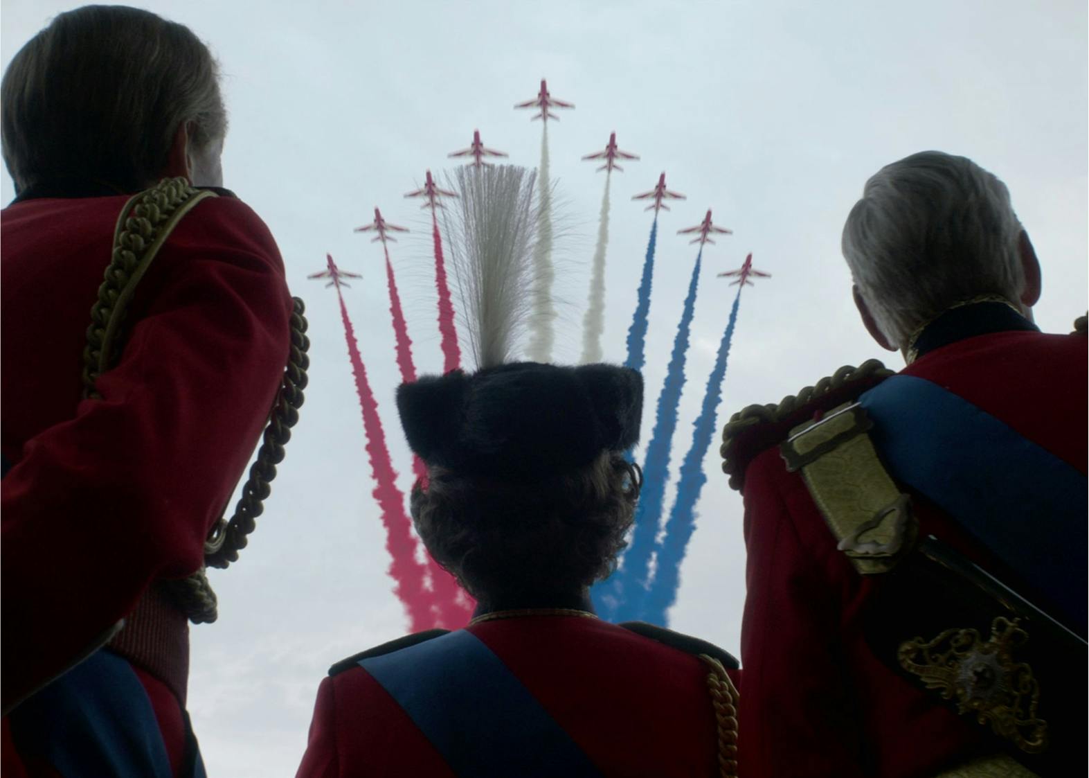 The silhouette of the Queen and two other men wear military garb and watch as planes shoot into the sky with red, white, and blue streams behind them.