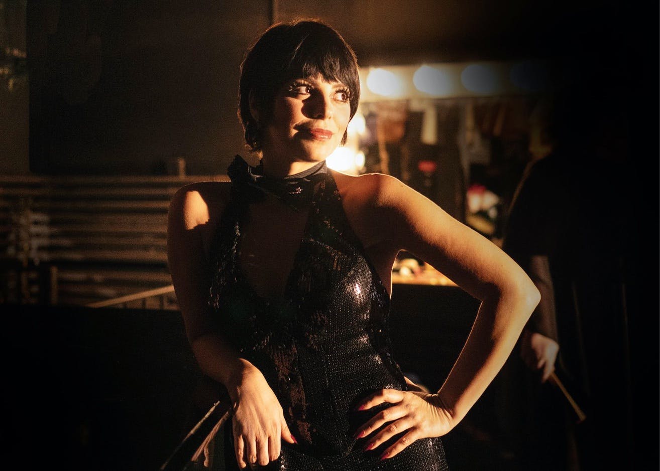 Krysta Rodriquez stands with her hand on her waist. The shot is mostly black, with light on her arm, face, and background. She wears a black sequiny dress and smiles at something in the distance.