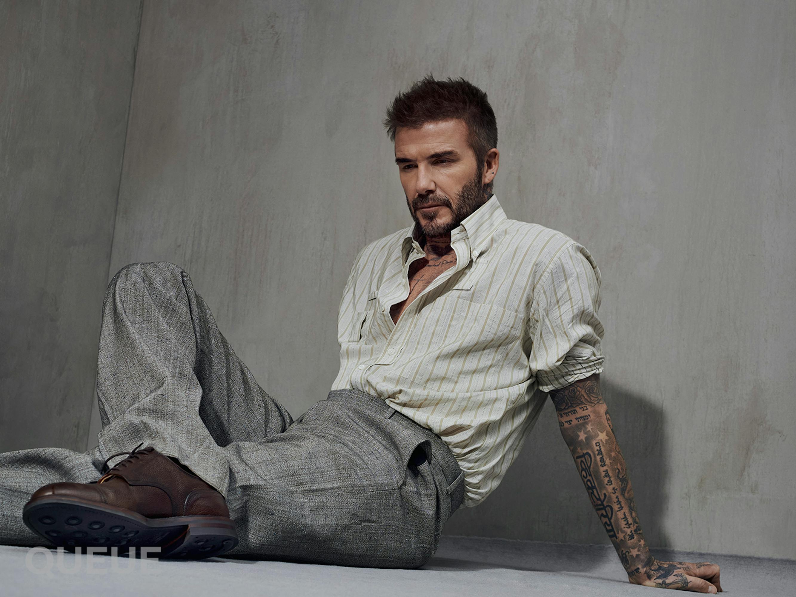 David Beckham wears gray pants and a light gray shirt and rests against a gray wall.