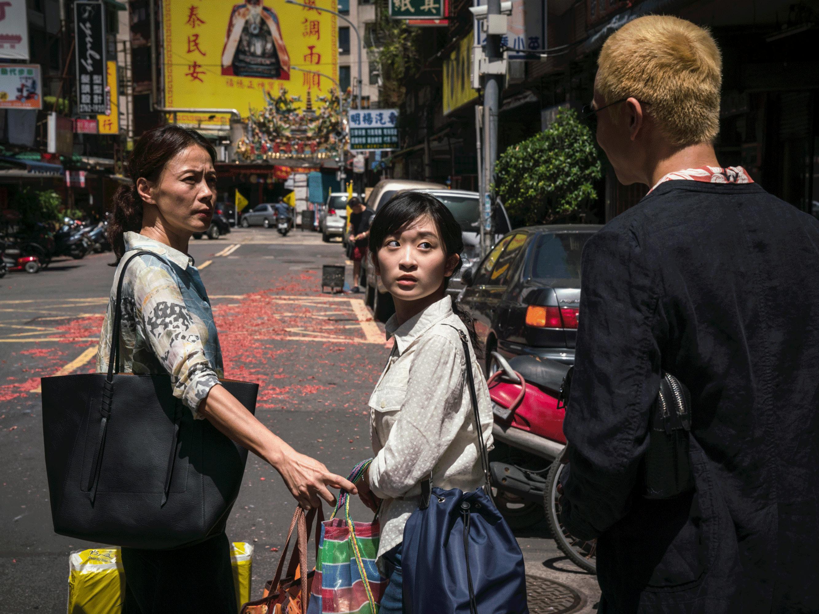 Miss Qin and Xiao-yu stop in the middle of a city street, arms laden with shopping bags, looking at Radish. Miss Qin reaches out an arm to Xiao-yu as if to usher her onward, while Xiao-yu gives Radish an expectant look. 