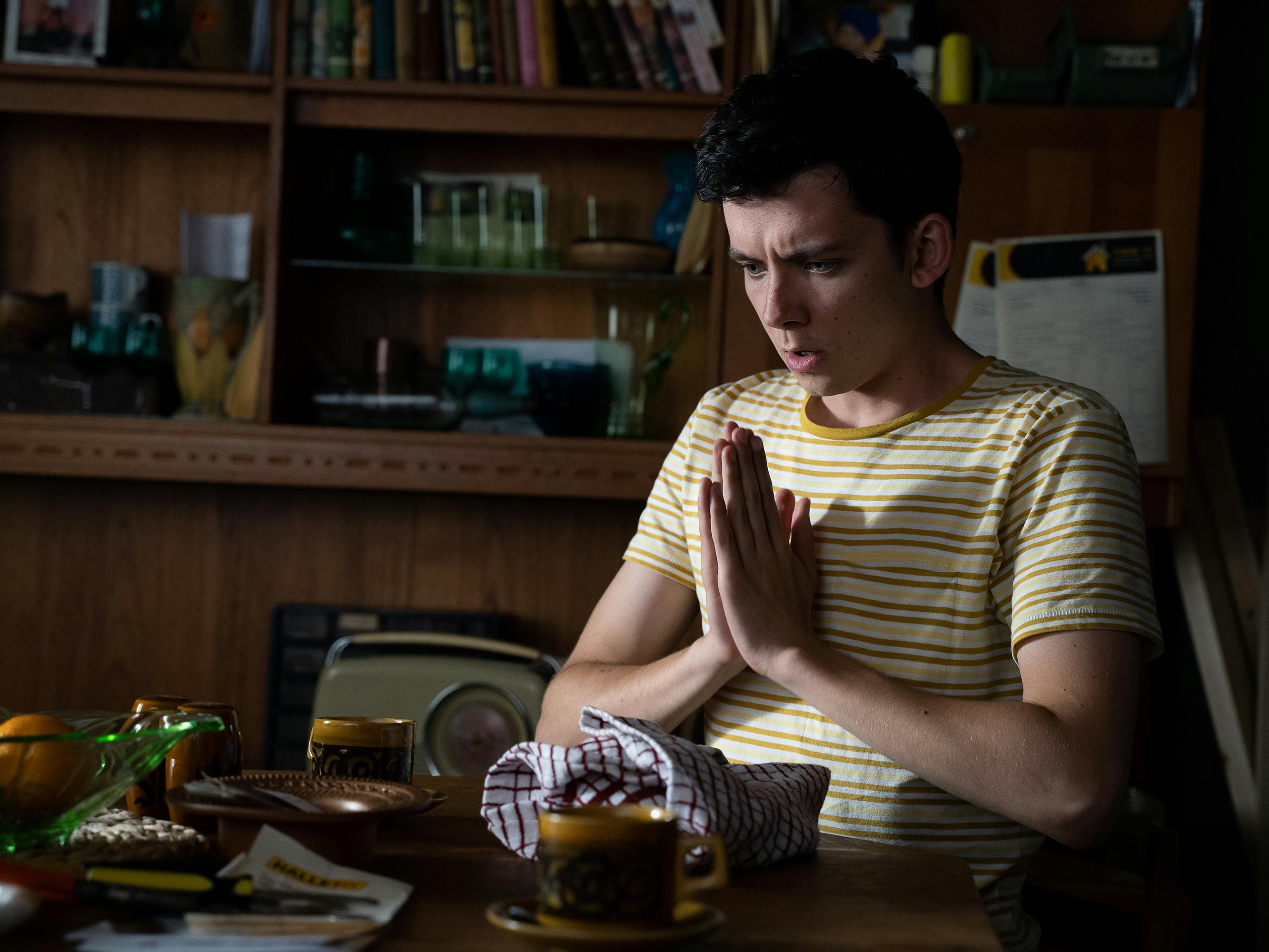 Otis Milburn (Asa Butterfield) wears a striped yellow shirt and looks like he is praying at a kitchen table in his eccentric home.