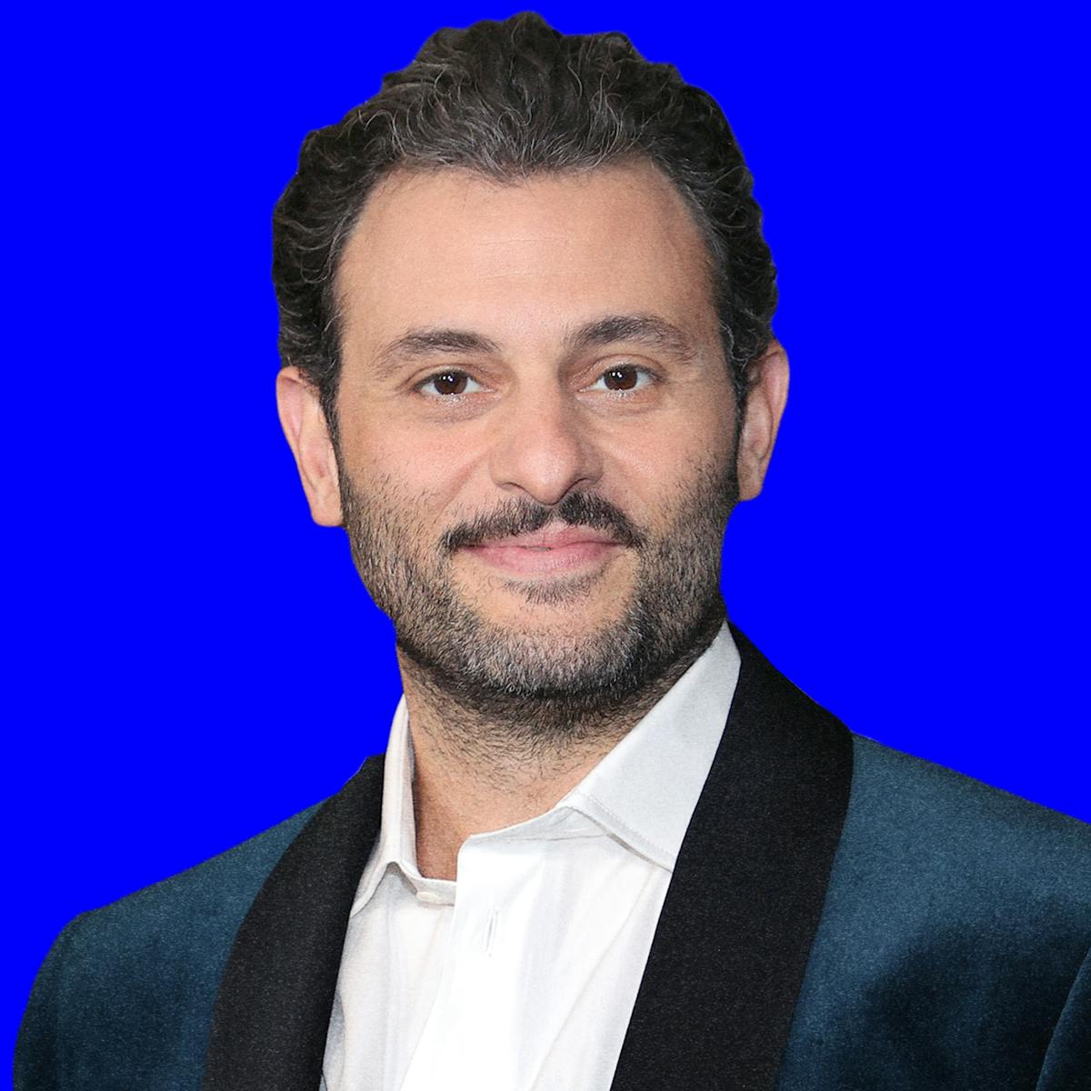 Arian Moayed wears a velvet jacket and a white buttoned down shirt. He has a short mustache and smiles at the camera. Behind him is a blue background with his name in capital letters repeated.