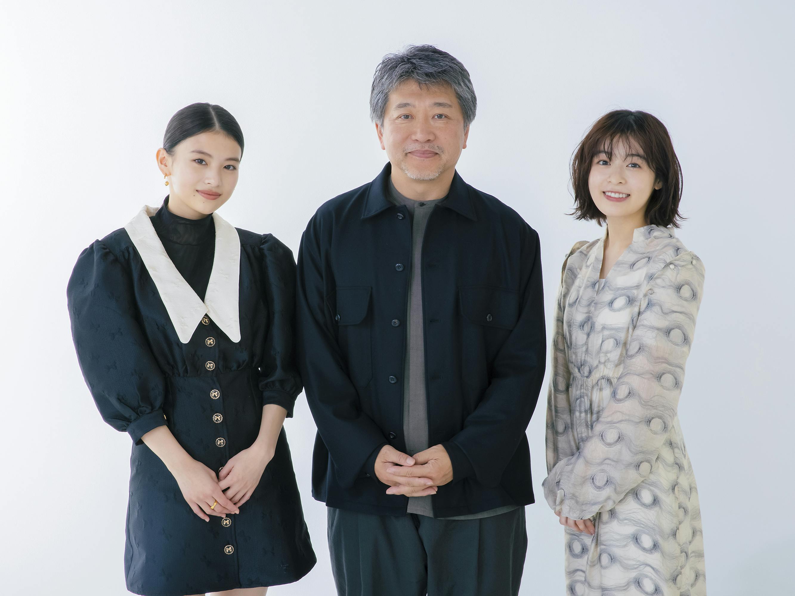 Natsuki Deguchi, Hirokazu Kore-eda, and Nana Mori stand together against a white background. Natsuki wears a black dress with a dramatic white color; Hirokazu wears a dark jacket and a grey t-shirt; Nana wear wear a grey-and-white dress.