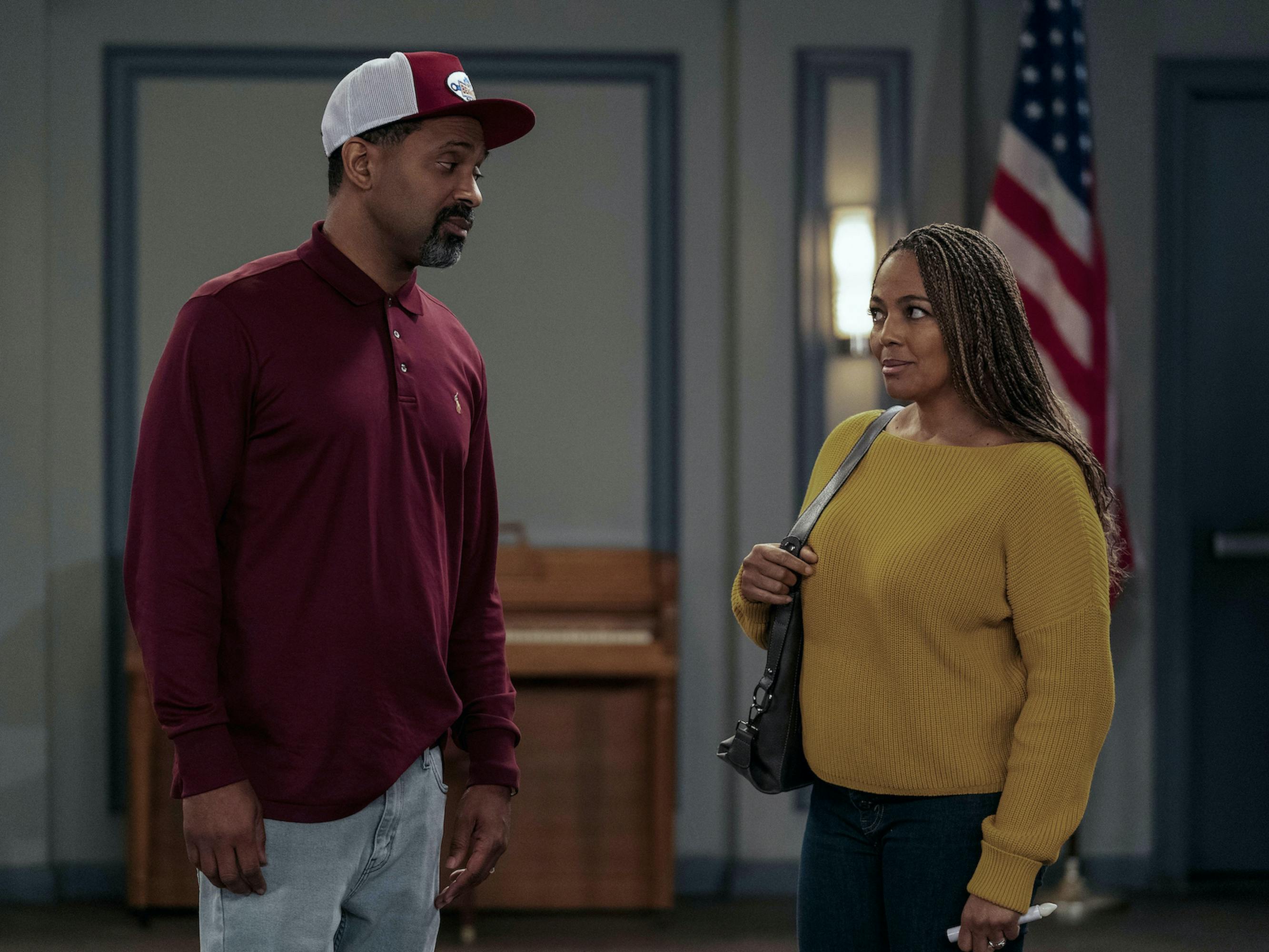 Mike Epps playing Bennie and Kim Fields playing Regina stand together. Epps wears a maroon shirt with a white and maroon hat. Fields wears a yellow sweater, carries a bag, and looks skeptically at Bennie. There is an American flag in the background.