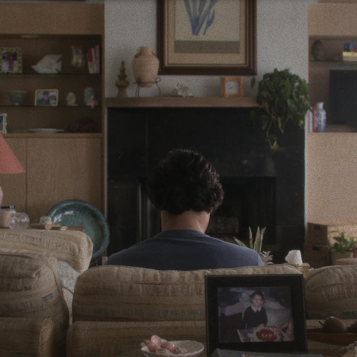 Joe (Charles Melton) sits on the living room couch reading in front of a dark TV screen.
