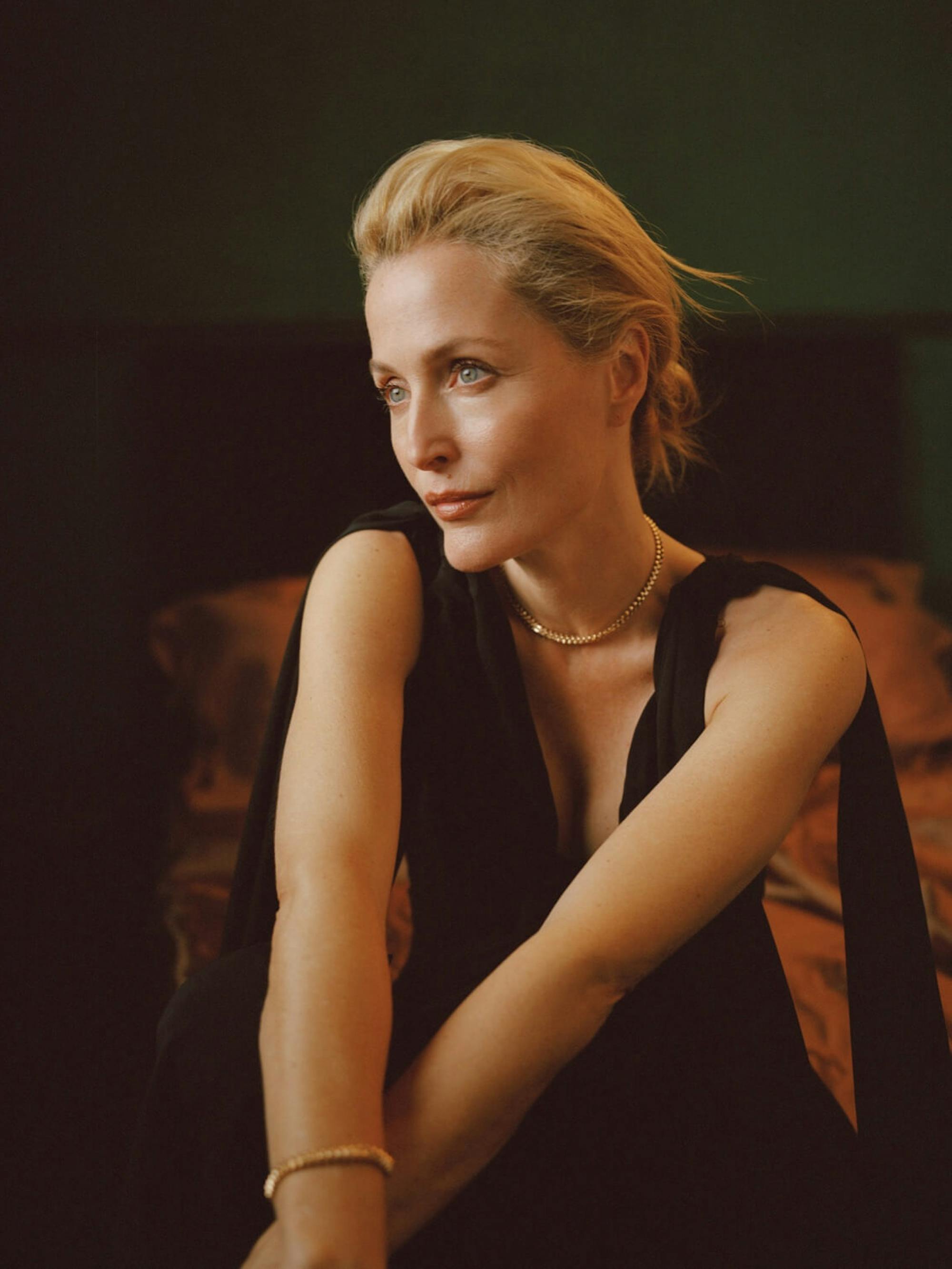 Gillian Anderson wears a black top and crosses her arms. Her wrists and neck are adorned with gold jewelry.
