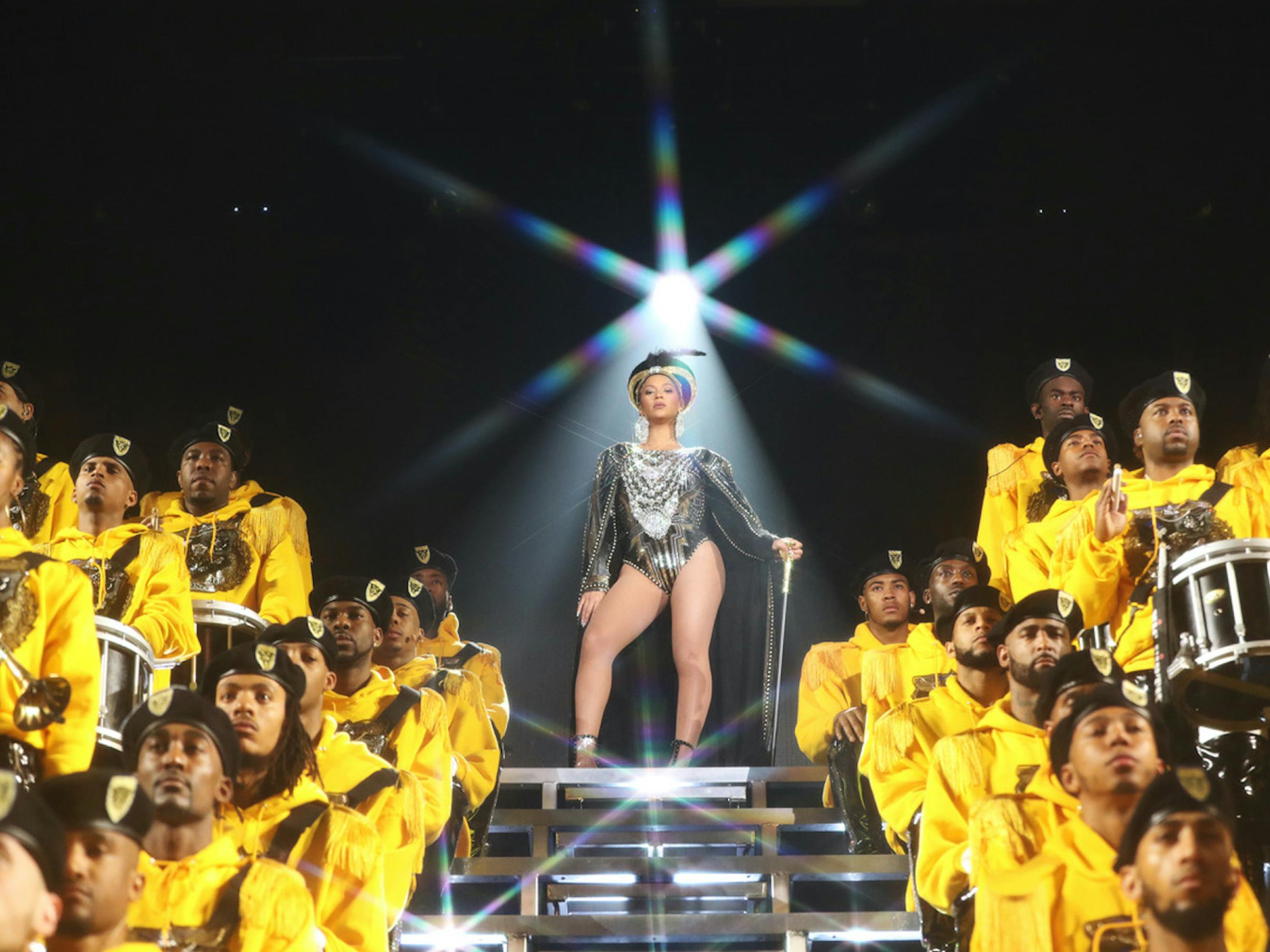 Beyoncé wears a silver leotard and stands amidst a marching band dressed in yellow.