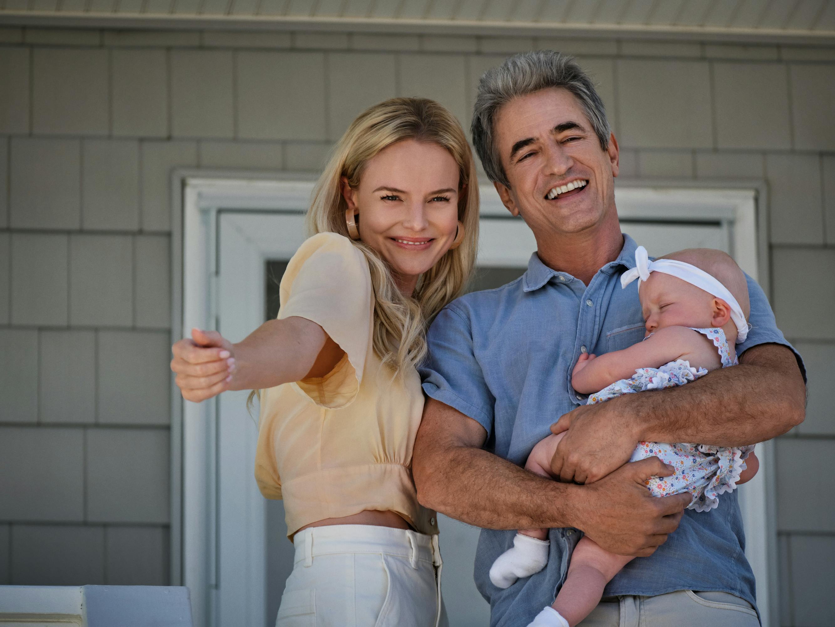 Heidi (Kate Bosworth) and Robert (Dermot Mulroney) stand on a porch holding a cute baby. She wears a yellow top and white pants, he wears a blue polo and khakis. The baby wears a white headband, a white onesize, and white socks.