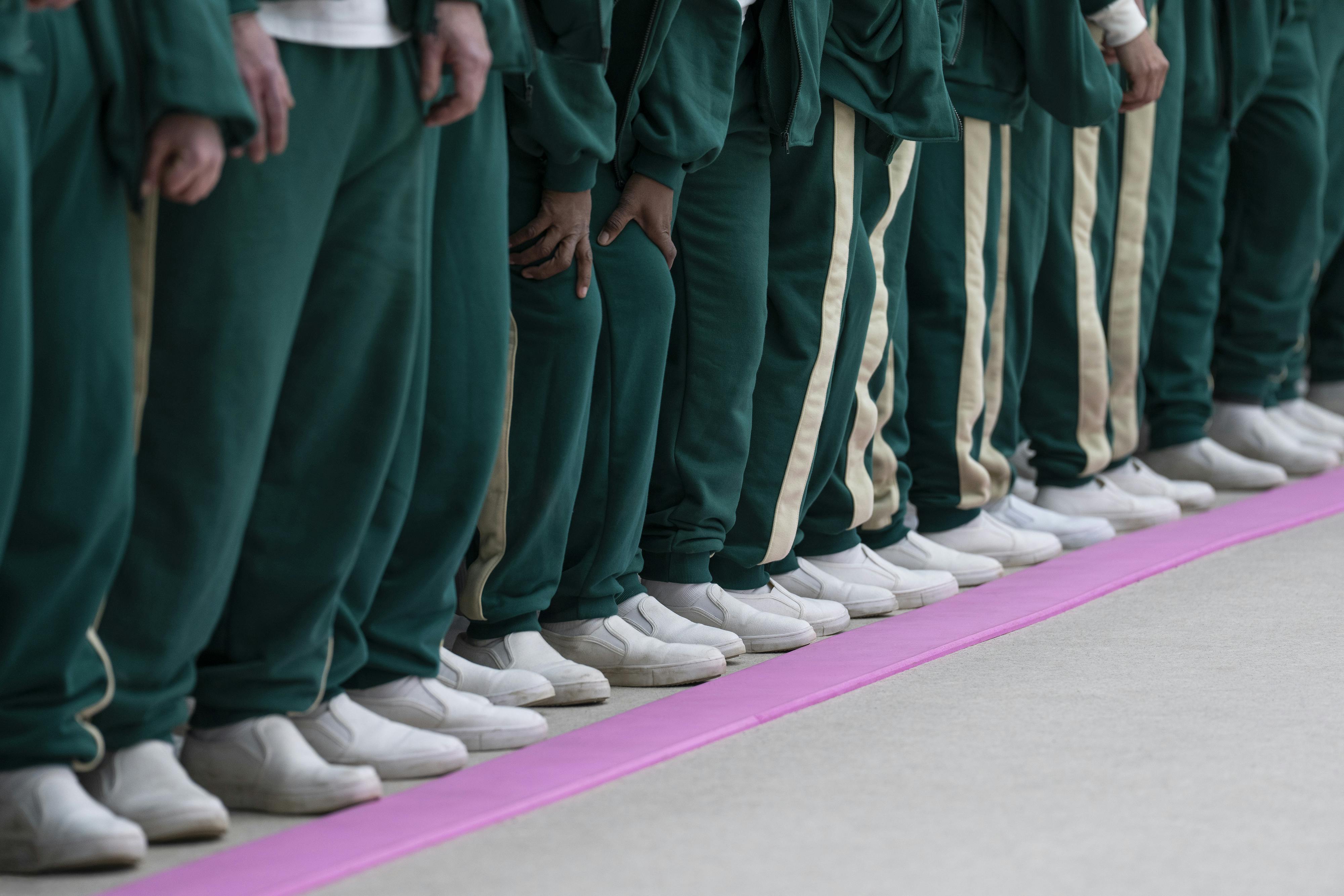 A bunch of people in green tracksuits and white shoes stand behind a pink line.