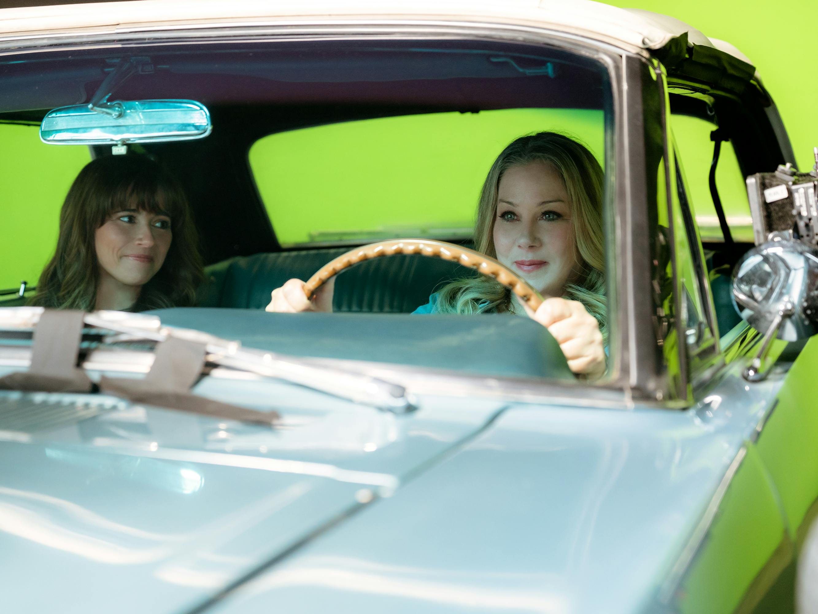 Linda Cardellini and Christina Applegate drive in a light blue car against a green screen. 